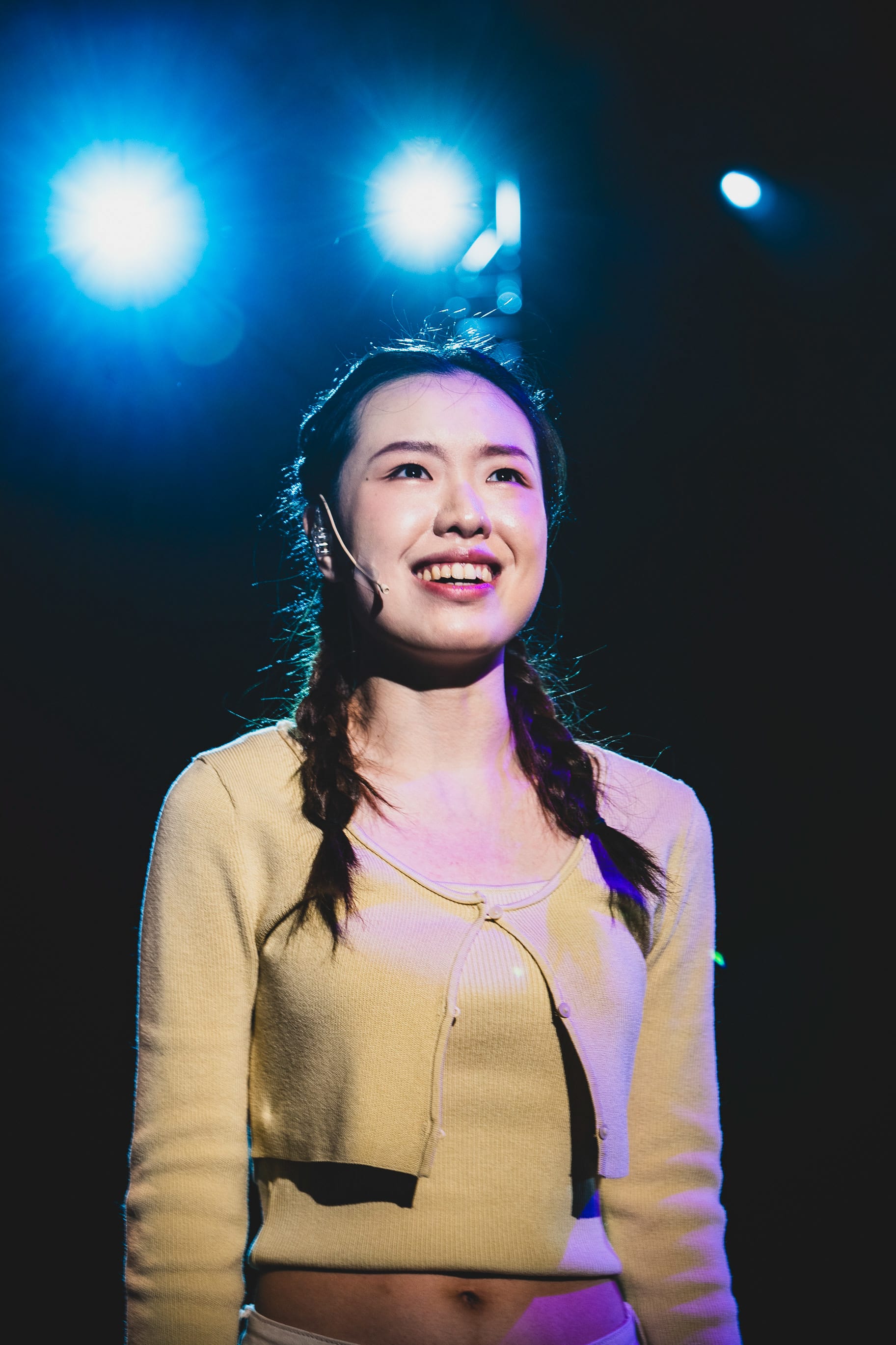 天台製作（Rooftop Productions）現代劇場作品《天真與世故之歌》的舞台劇照，沈卓瑤在藍色舞台燈光下演唱，她紮著雙辮，身穿淺黃色開衫，面露燦爛笑容。這張於西九龍文化區拍攝的劇照捕捉了她投入演繹角色的一刻，感嘆道：「I wish that I could turn back time」