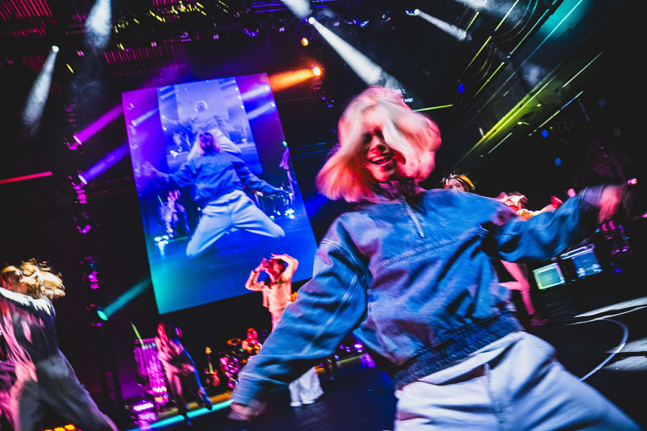 A dynamic performance shot from Rooftop Productions' contemporary theatre piece 'Songs of Innocence and Experience' at the West Kowloon Cultural District. In the foreground, a dancer with blonde hair is captured in motion wearing a denim jacket, while in the background, a large screen displays a projection of performer Wu Lui-fung (Beelzebub) in mid-air. The stage is bathed in dramatic, vibrant purple and gold lighting, emphasizing the raw energy of this Hong Kong stage production. The performer on screen echoes the high-energy performance with the line: 'HEYYEYAAEYAAAEYAEYAA'