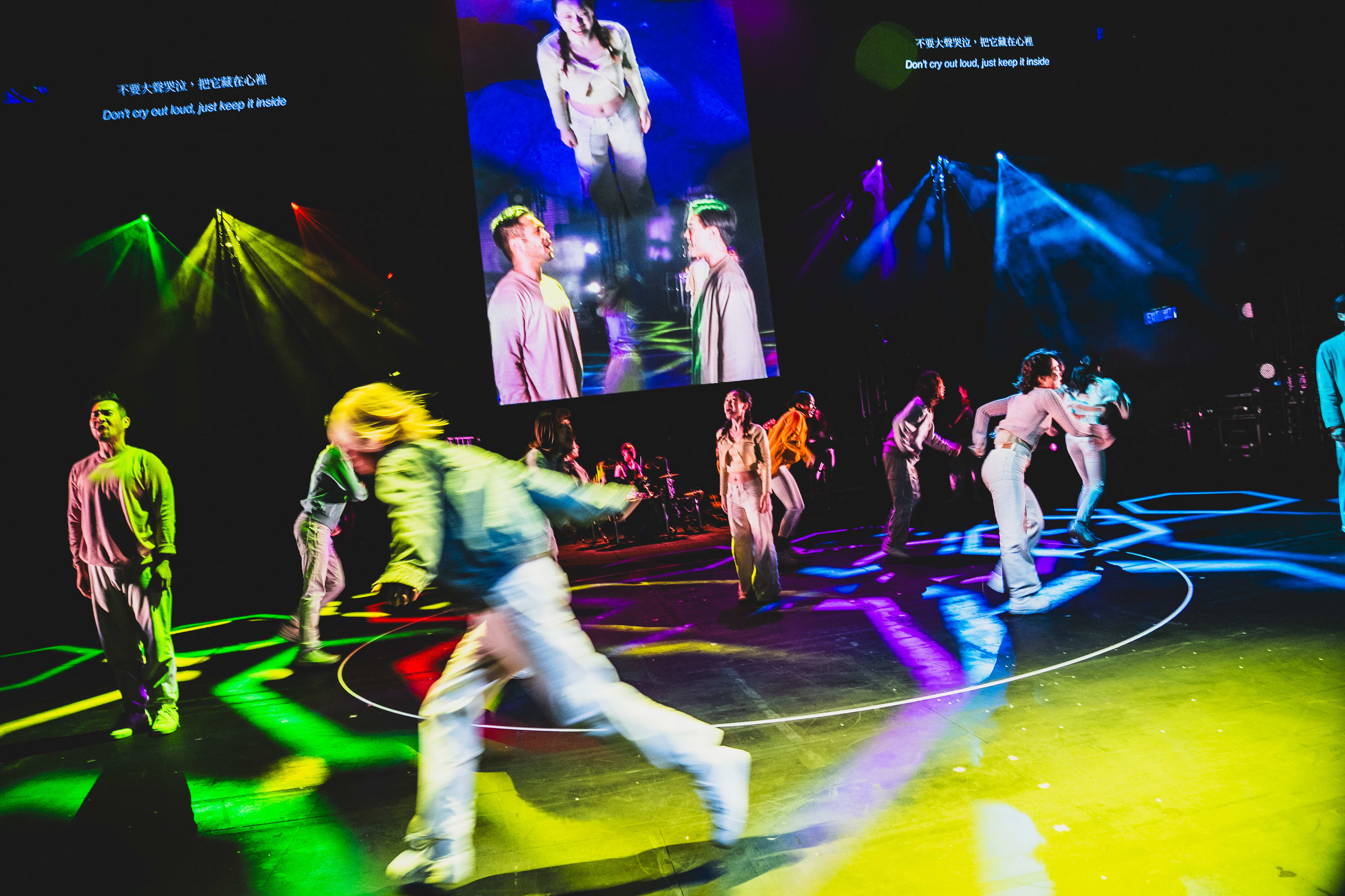 A dynamic wide shot of the contemporary dance-theatre performance 'Songs of Innocence and Experience' at the West Kowloon Cultural District, featuring actors including Sum Cheuk-yiu (Abdiel) moving across a dark stage illuminated by vibrant green, yellow, and blue spotlights. A large digital screen in the background projects close-up footage of the performers, while the stage floor is marked by circular lighting patterns. Sum Cheuk-yiu is captured mid-movement, epitomizing the emotional intensity of this Rooftop Productions show. As the performers express the internal struggle of the narrative, they sing, 'Don't cry out loud, just keep it inside'.