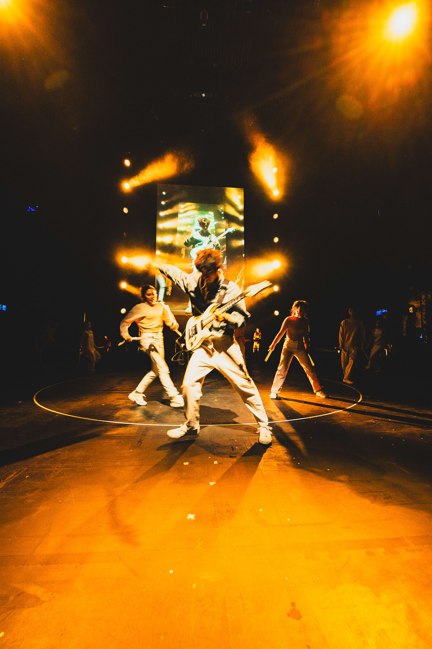 A dramatic stage shot from Rooftop Productions’ contemporary theatre piece 'Songs of Innocence and Experience' at the West Kowloon Cultural District. The image features performer Wu Lui-fung center stage, energetically playing a guitar while embodying the character Beelzebub. He is surrounded by other cast members in a ring of light, with warm, intense amber stage lighting highlighting their silhouettes. The backdrop features a digital screen showcasing artistic visuals, capturing a high-energy musical moment from the production. 'Digimon solo'