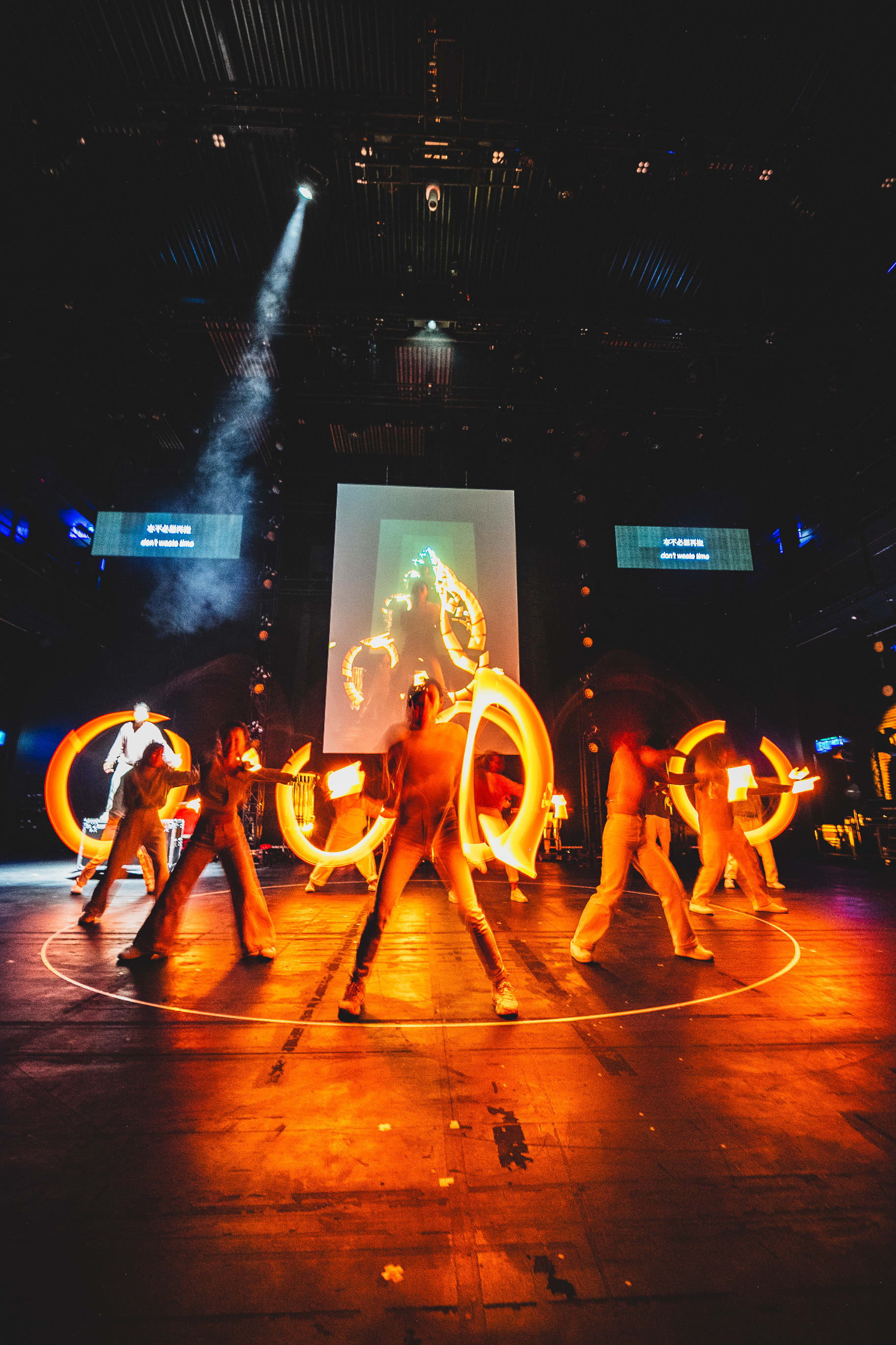 A dramatic wide shot from Rooftop Productions’ contemporary theatre piece 'Songs of Innocence and Experience' (《天真與世故之歌》) at West Kowloon, capturing a powerful ensemble performance on stage. Dancers are positioned within a glowing circular light on the floor, holding circular light tubes that create blurred trails of motion, emphasizing energy and defiance. The stage is bathed in deep amber and orange hues, with a large screen displaying abstract visuals in the background and a spotlight cutting through the darkness. The scene culminates in a fierce stance as the cast echoes the line, 'Oh, let's fight.'