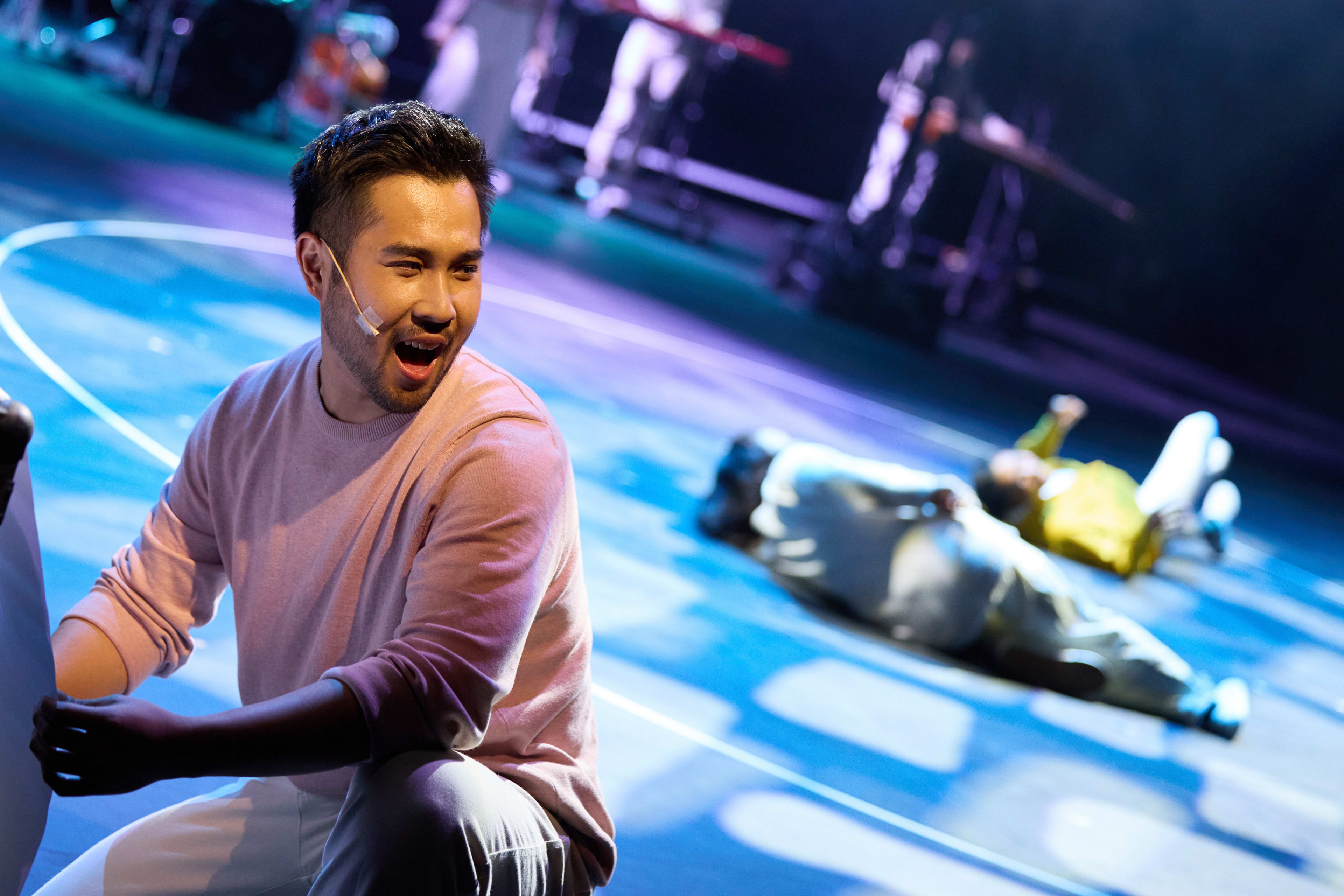 In a dramatic scene from Rooftop Productions' contemporary theatre piece 'Songs of Innocence and Experience' at the West Kowloon Cultural District, Mok Kok-pong (as Michael) is captured in a dynamic moment on stage. Kneeling in the foreground, he wears a light pink sweater and looks toward the audience with an intense, expressive gaze, wearing a small headset microphone. Behind him, under vibrant blue stage lighting, another performer lies on the floor, creating a sense of emotional depth and theatrical tension. 'Opening titles'