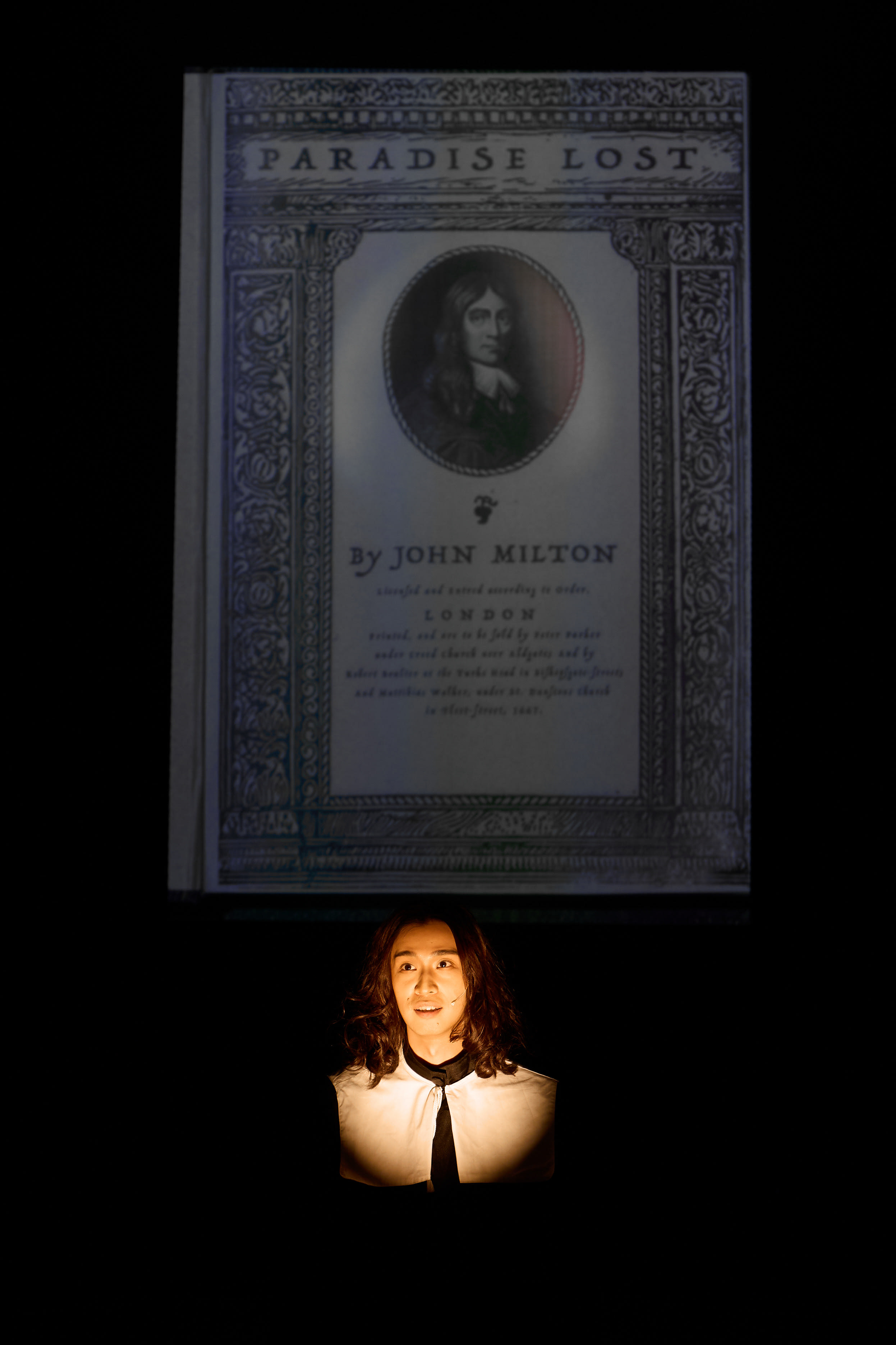 A dramatic stage shot from Rooftop Productions' contemporary theatre performance, 'Songs of Innocence and Experience' (《天真與世故之歌》). Actor Mak Ho-tin is illuminated by a sharp spotlight against a dark background, embodying the poet John Milton. Behind him, a large projection of the title page of John Milton's 'Paradise Lost' looms, creating a striking visual juxtaposition between the performer and the literary work. John Milton.