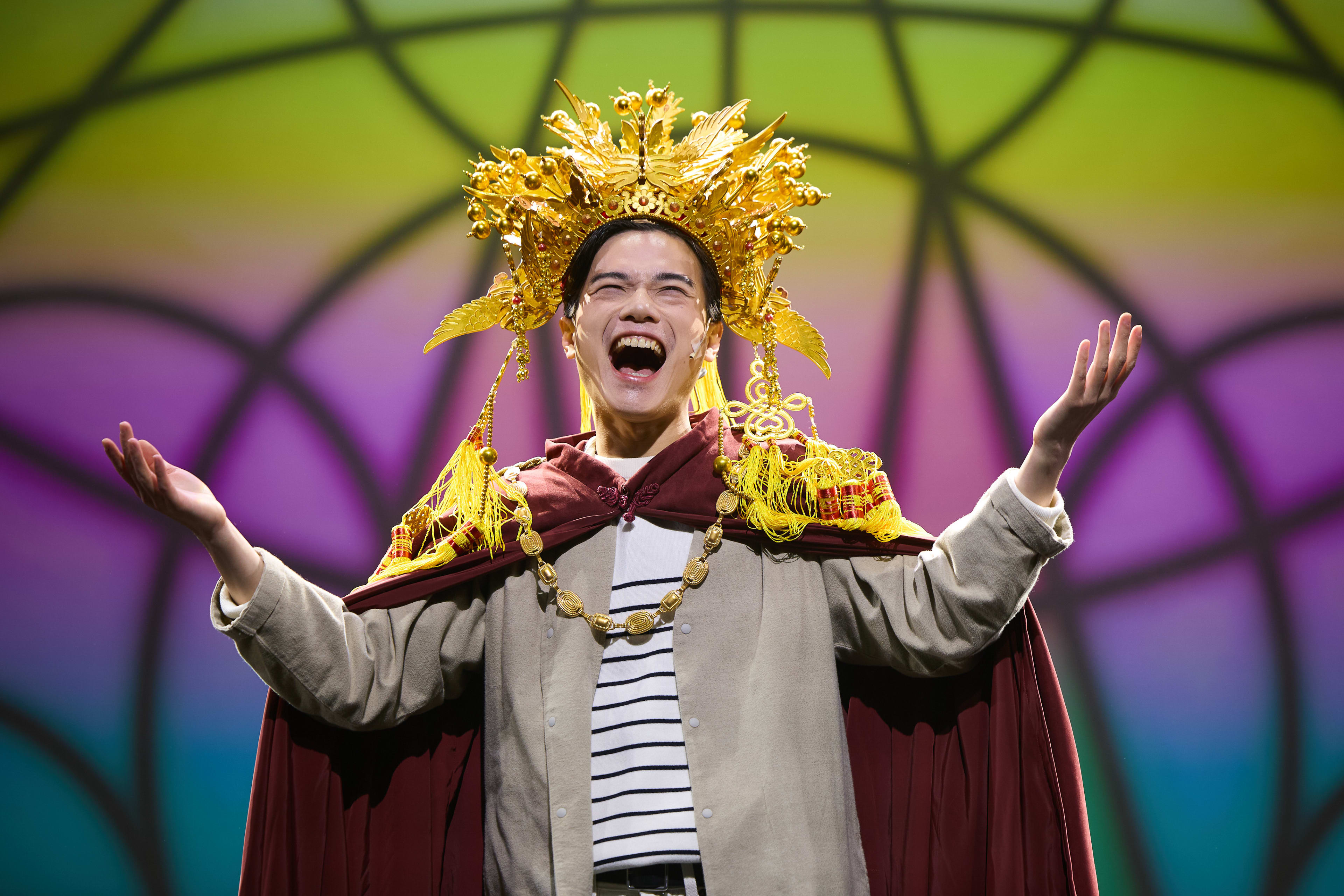 Lo Man-chak performs as God in Rooftop Productions' contemporary theatre piece 'Songs of Innocence and Experience'. He wears a grand, golden, intricate headpiece with dangling tassels and a deep maroon cape over a casual striped top, his arms outstretched in a dramatic, expressive gesture under vibrant, multicoloured stage lighting. God addresses the angels.