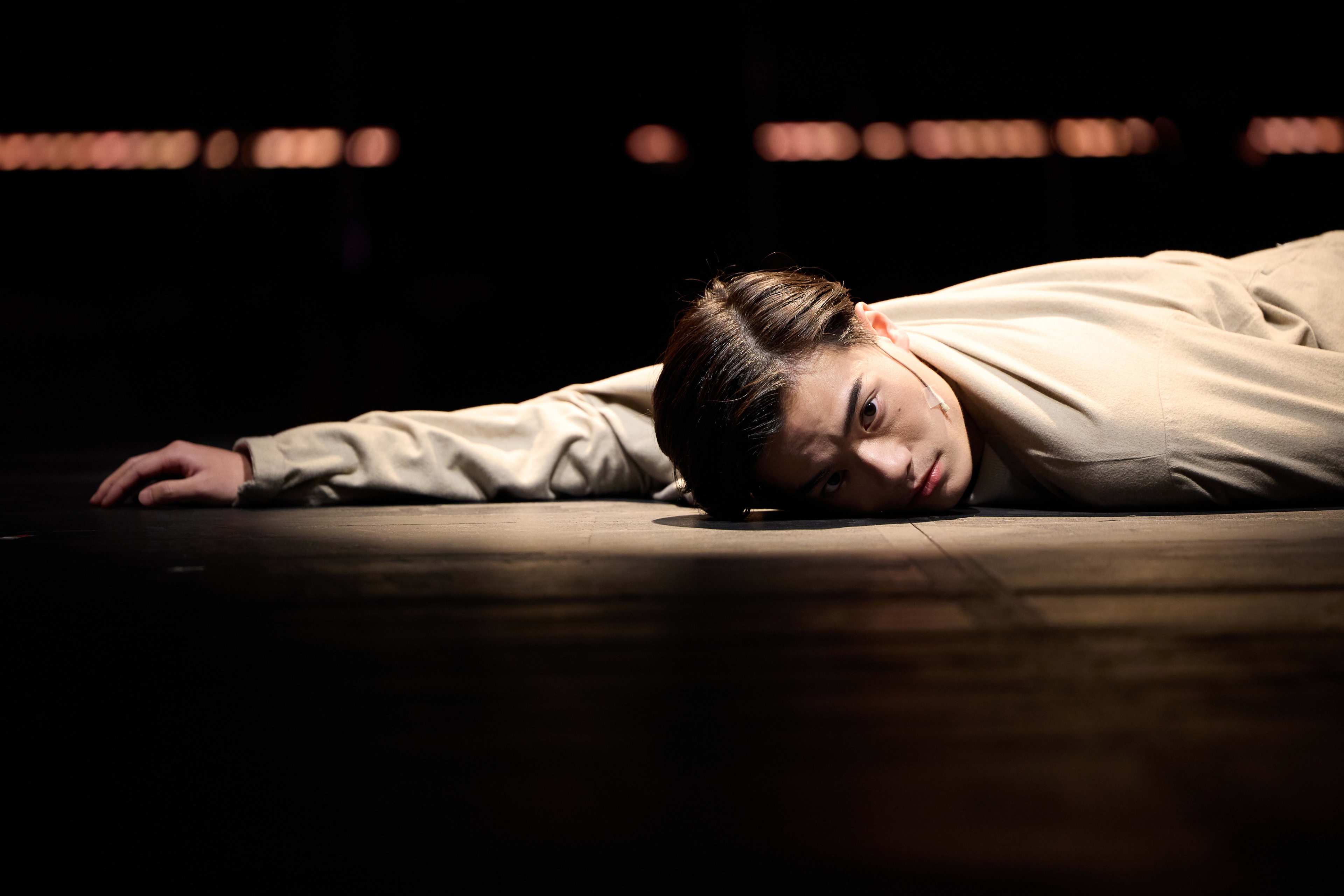 In a dramatic scene from Rooftop Productions' contemporary theatre piece 'Songs of Innocence and Experience' (《天真與世故之歌》), actor Lo Man-chak lies prone on the dimly lit wooden stage. Bathed in a focused spotlight against a dark background, his expression is one of vulnerability and introspective stillness, capturing the intensity of a live Hong Kong stage performance. 'Confession'
