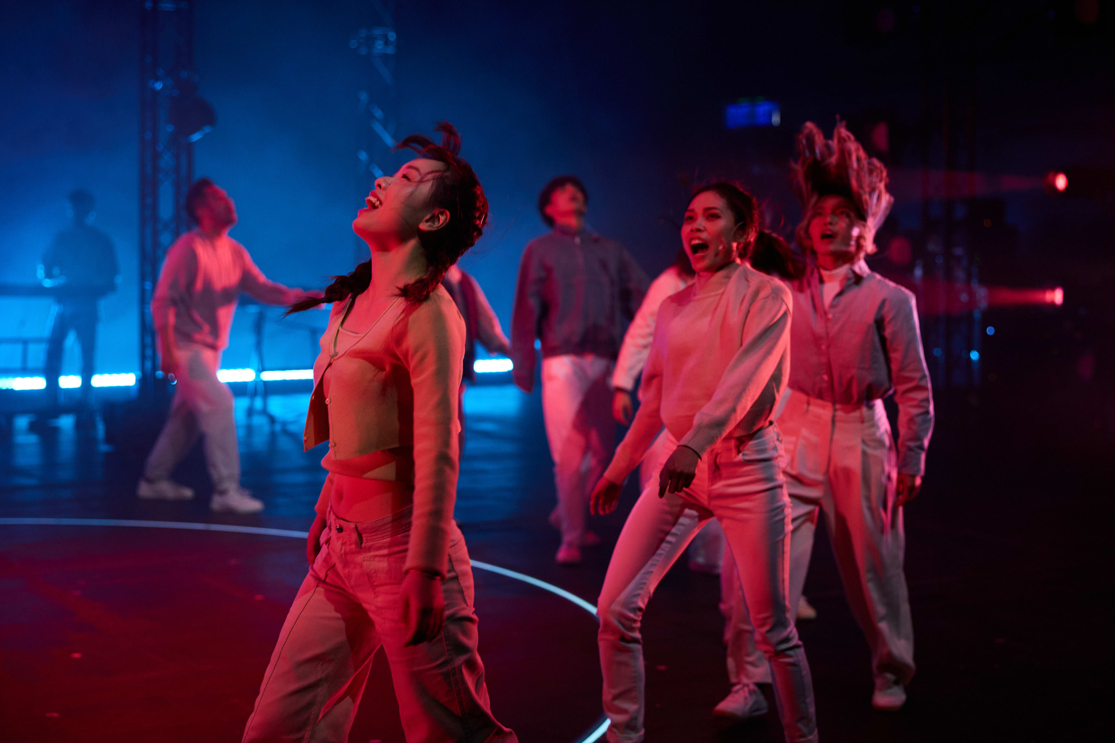 A dramatic stage shot from Rooftop Productions' contemporary theatre performance, 'Songs of Innocence and Experience' (《天真與世故之歌》), set in the West Kowloon Cultural District. On a dimly lit stage defined by circular floor lighting, a group of performers, including Sin Lok-yan and Chin Wing-kar, are captured in an intense moment of movement. Sin Lok-yan, portraying Eve, takes center stage with her head tilted back in an expression of raw emotion, while other cast members move dynamically around her under moody blue and crimson spotlights, capturing the tension of the scene. The performers seem to be 'Waiting in hell'.