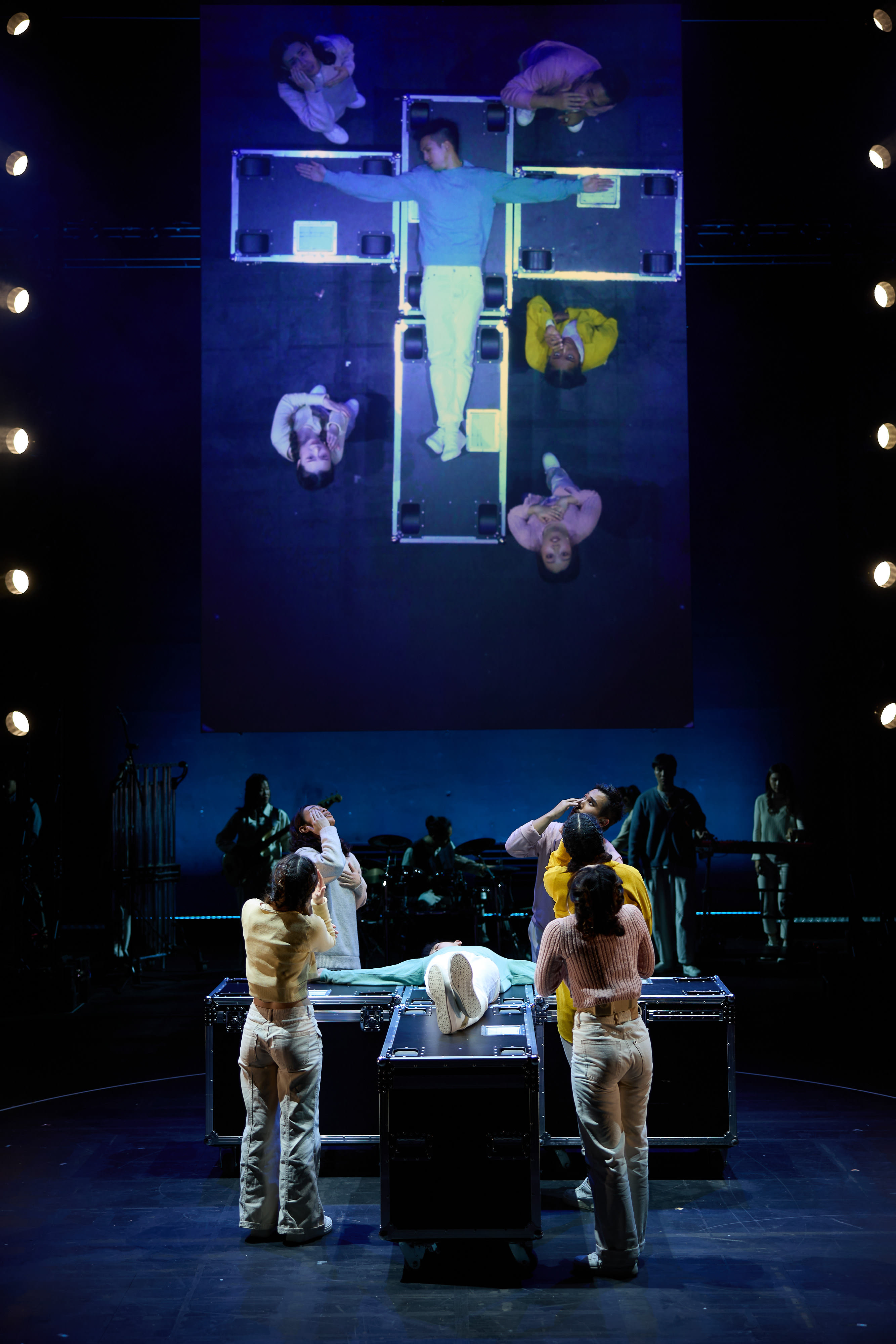 In this dramatic scene from Rooftop Productions' contemporary theatre piece 'Songs of Innocence and Experience' (《天真與世故之歌》), performers stand on a dimly lit stage in front of a large, vertical screen. The screen projects a top-down, bird's-eye view of actors lying on flight cases arranged in a cross formation. On stage, performers in casual attire cluster around similar flight cases, interacting with a central figure who lies flat, mirroring the composition on the projection. The moody, stark lighting emphasizes the thematic tension between spiritual motifs and modern theatrical art. A performer standing by the center console softly delivers the line: 'But I'm not Jesus...'