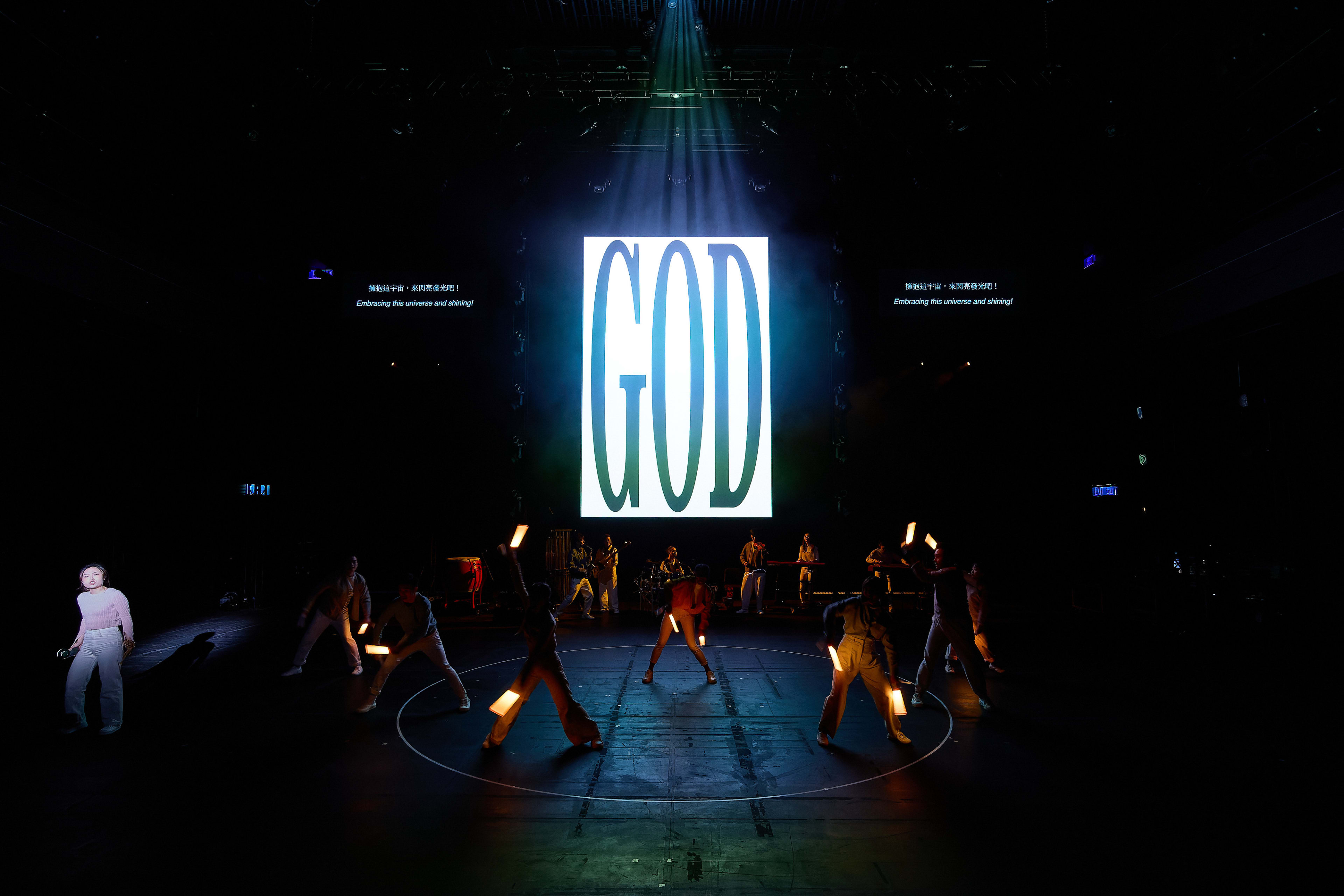 In a dramatic scene from Rooftop Productions' contemporary theatre performance 'Songs of Innocence and Experience' at the West Kowloon Cultural District, a large screen featuring the word 'GOD' in bold, vertical typography looms over a dark stage. A performer, Lee On-sang (as Moloch), stands alone in a spotlight on the left, while a group of dancers in silhouette move in a circular formation, holding glowing light wands that cut through the darkness. The scene captures a powerful tension between light and shadow. As the ensemble moves, Lee On-sang recites, 'God, Angels, Adam... A cruel angel's thesis.'