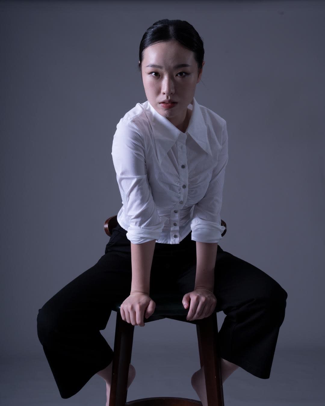 演員沈卓瑤為天台製作 (Rooftop Productions) 舞臺演出拍攝的宣傳照。照片中，她以充滿張力的姿態前傾，雙手撐在木椅上，眼神堅定銳利。她身著簡潔的白襯衫與黑長褲，背景為中性灰調，展現了當代香港劇場的極簡美學風格。