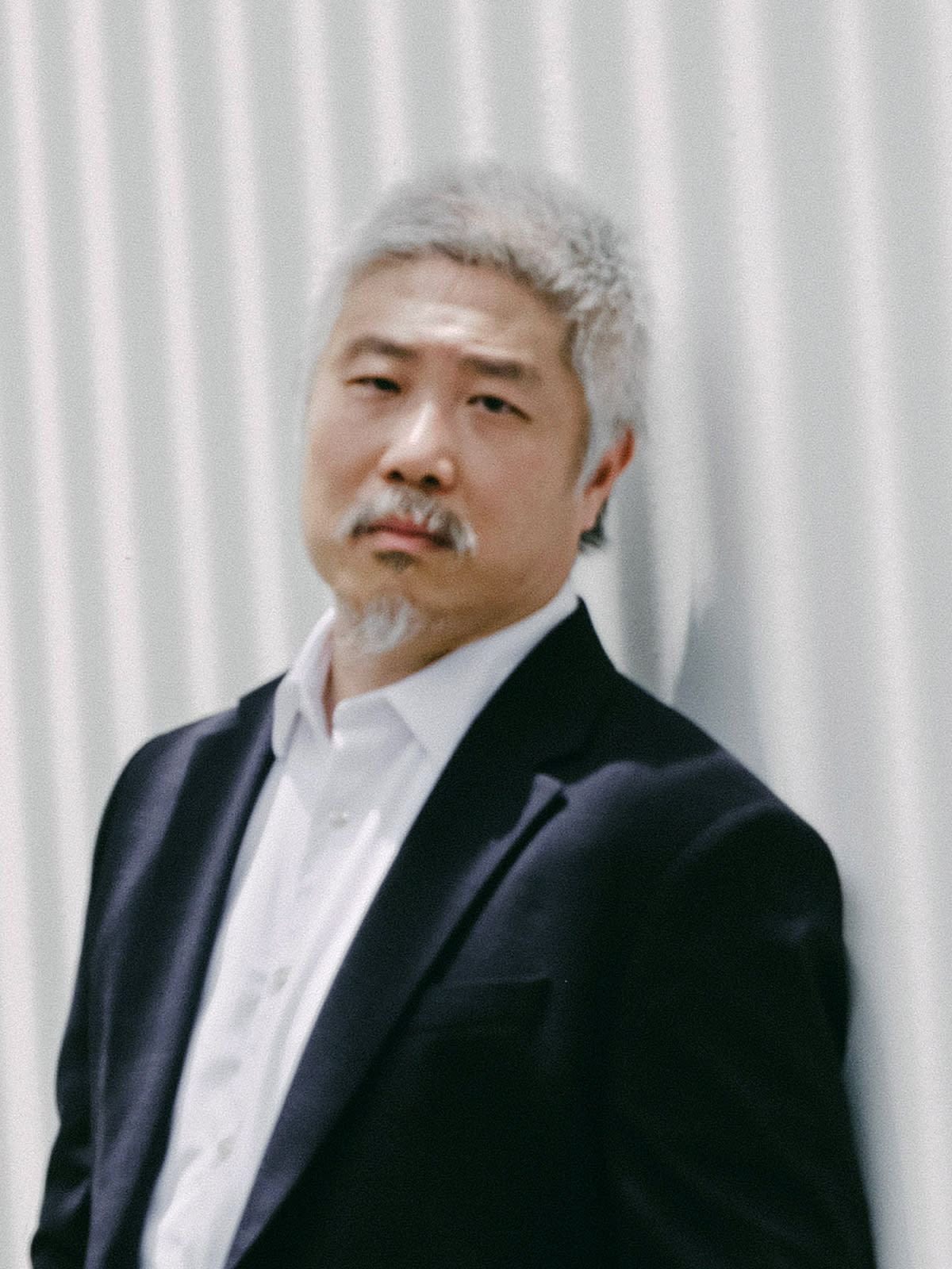 A professional headshot of Hong Kong theatre actor Sung Boon Ho, captured for Rooftop Productions. He stands against a clean, textured white background, wearing a dark blazer over a crisp white button-down shirt. His expression is serious and composed, reflecting the dramatic intensity often found in contemporary Hong Kong stage performances.