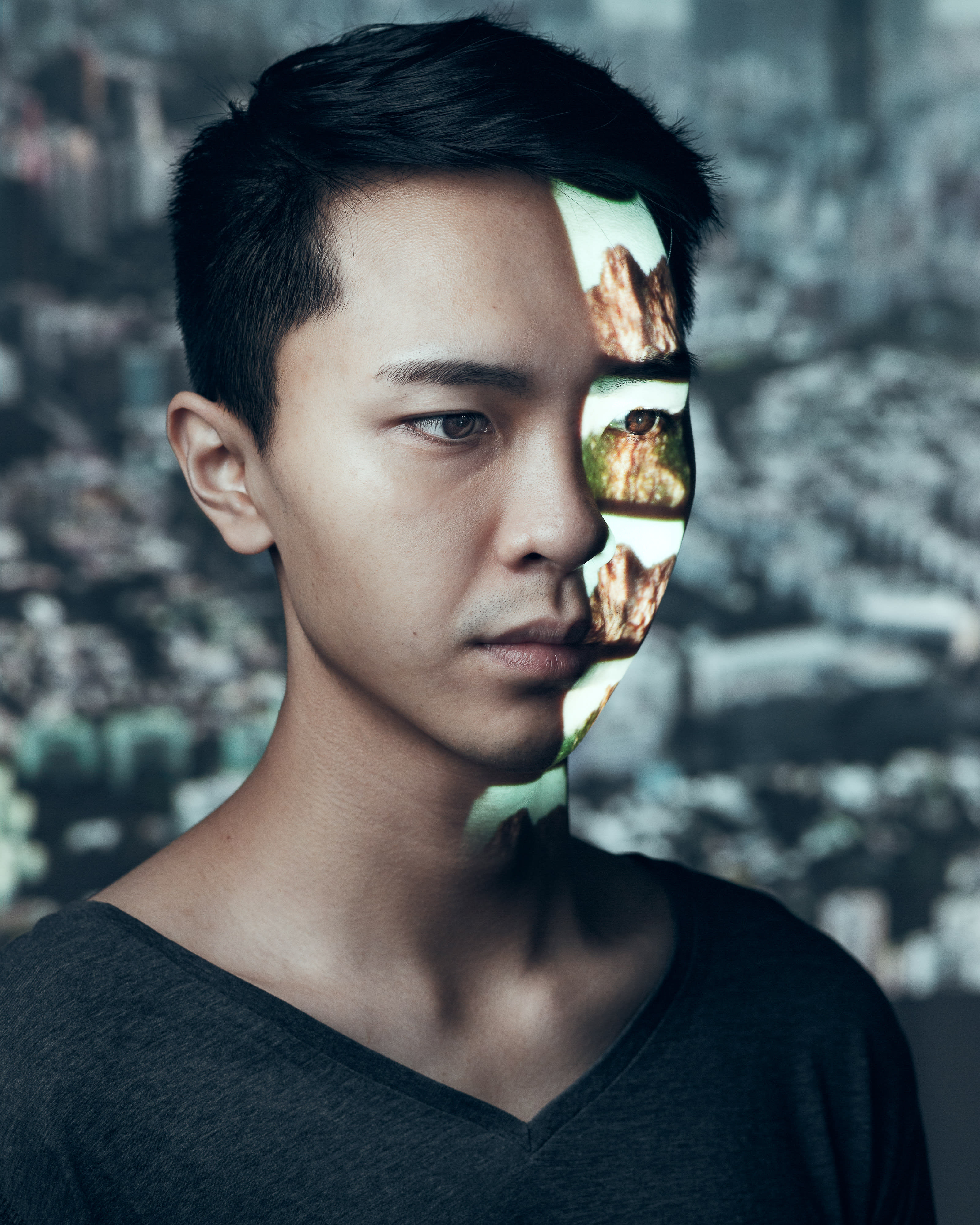 A dramatic headshot of actor Ng King Lung for the Rooftop Productions contemporary theatre piece 'Testimony' ('山下的證詞'). A projection of a rugged, mountainous landscape is cast onto the left side of his face, creating a striking contrast between his contemplative expression and the harsh, natural textures of the projected terrain, set against a blurred urban Hong Kong backdrop. 'Testimony'