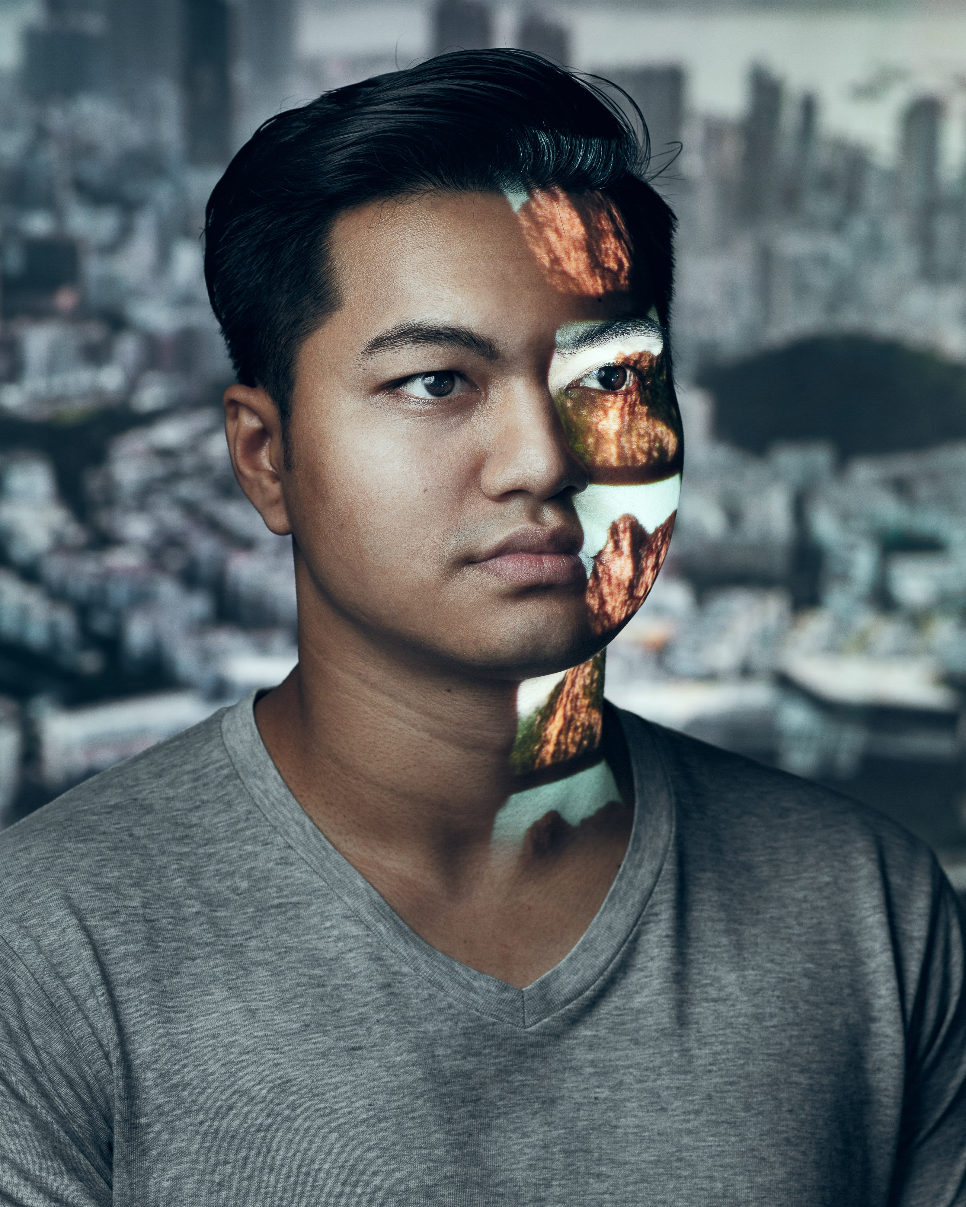 A dramatic promotional portrait for the contemporary Hong Kong theatre production Testimony, featuring actor Basnet Tusar. He is shown from the chest up against a blurred urban background, with a striking projection of rugged mountain textures cast across the right side of his face and neck, symbolising the collision of nature and memory. Testimony.