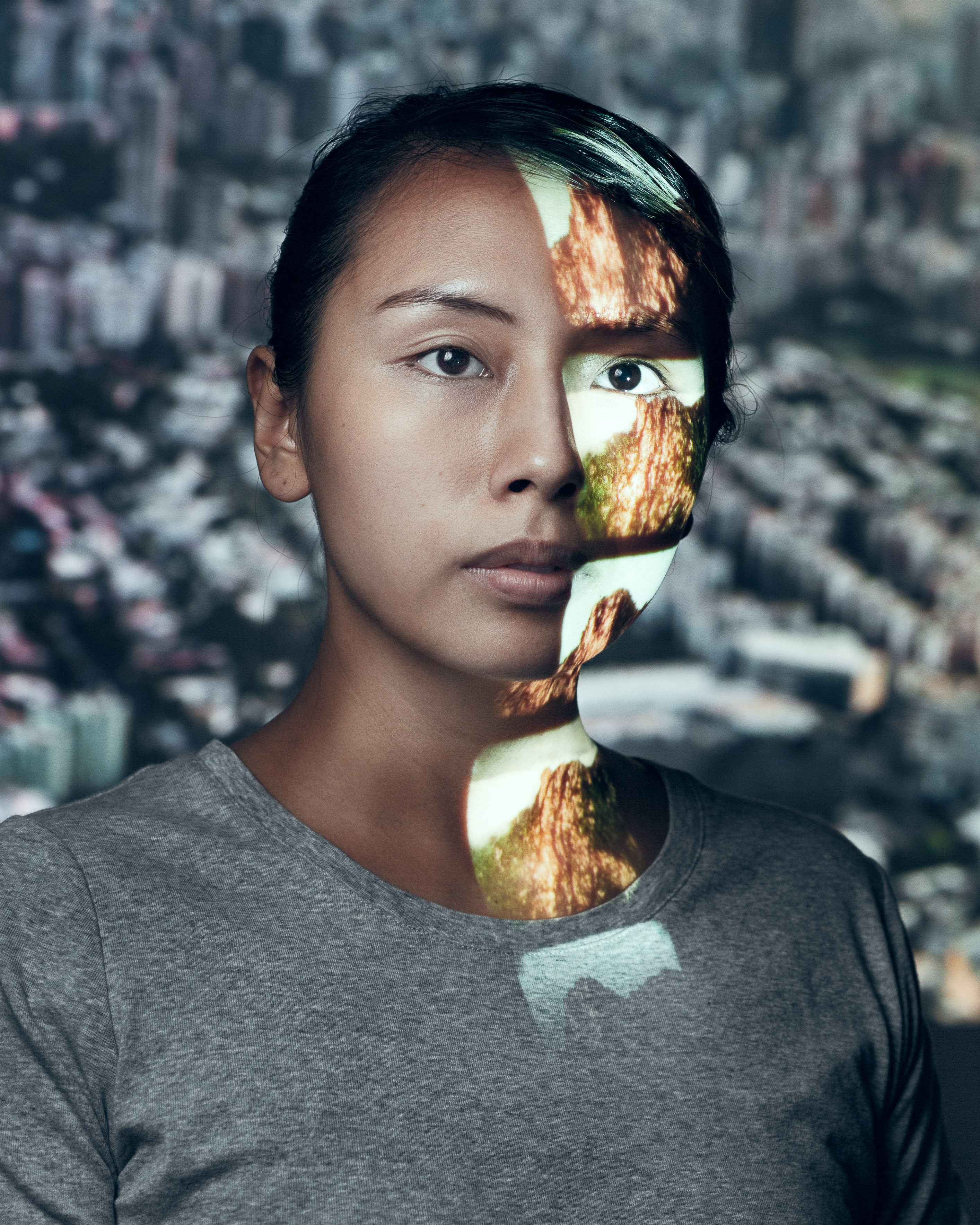 Portrait of performer Arisa Yasaratne for the Rooftop Productions contemporary theatre piece 'Testimony'. Her face is partially lit by a projection showing the rocky, rugged surface of a mountain, contrasting with the blurred Hong Kong cityscape in the background. Her expression is solemn and contemplative, highlighting themes of urban identity and memory in Hong Kong stage performance. "Testimony"