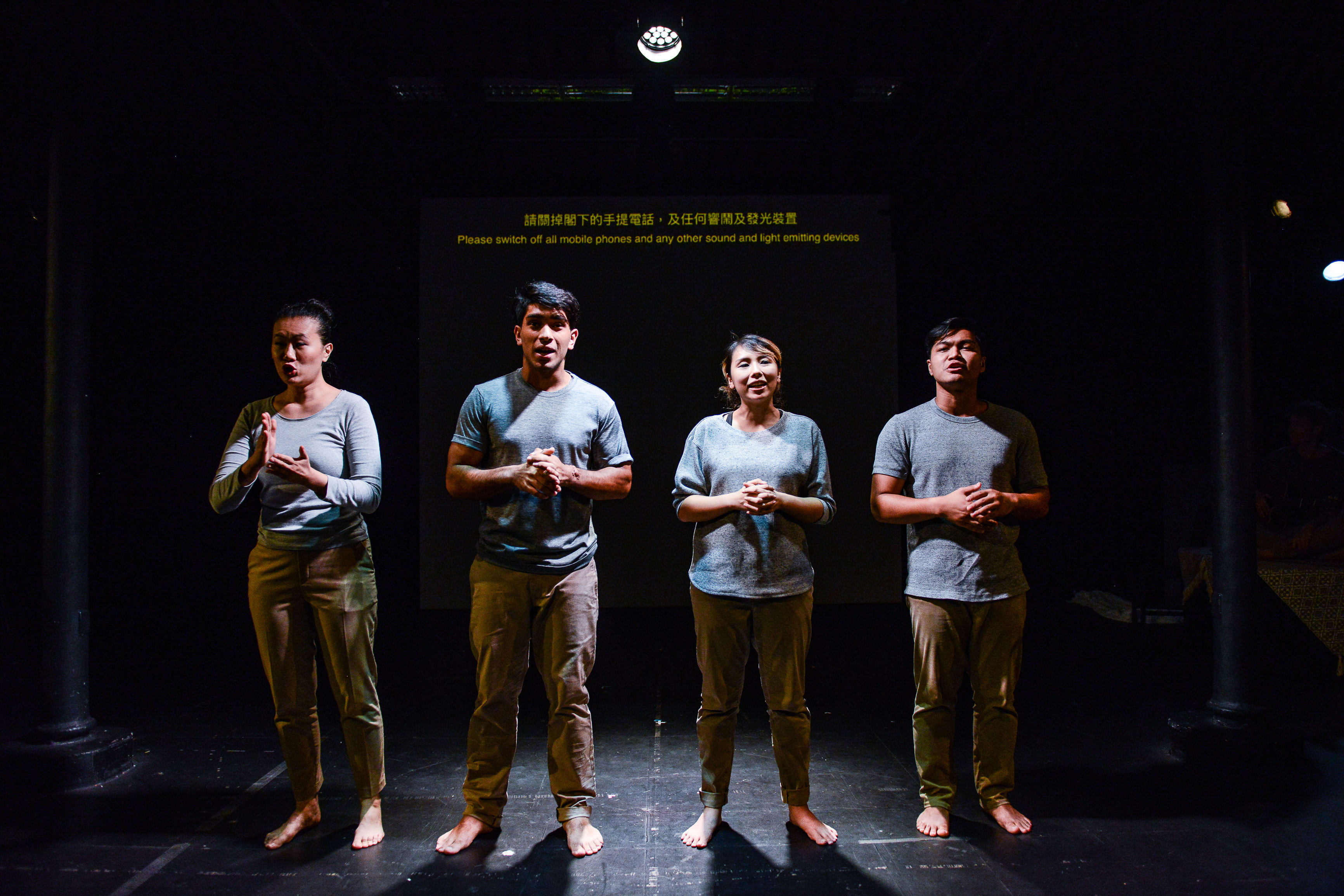 四位演員在香港當代劇場《山下的證詞》排練現場，並排站立於舞台中央。他們身穿休閒大地色系服裝，在頂部聚光燈的照射下，展現出極簡而充滿張力的戲劇氛圍。演員們（左起：李婉晶、Basnet Tusar、李里莎、巫加沙）表情嚴肅凝重，深刻詮釋出劇中沉重而發人深省的主題。他們身後的螢幕模糊地顯示著中英文提示，提醒觀眾關閉電子設備。《山下的證詞》