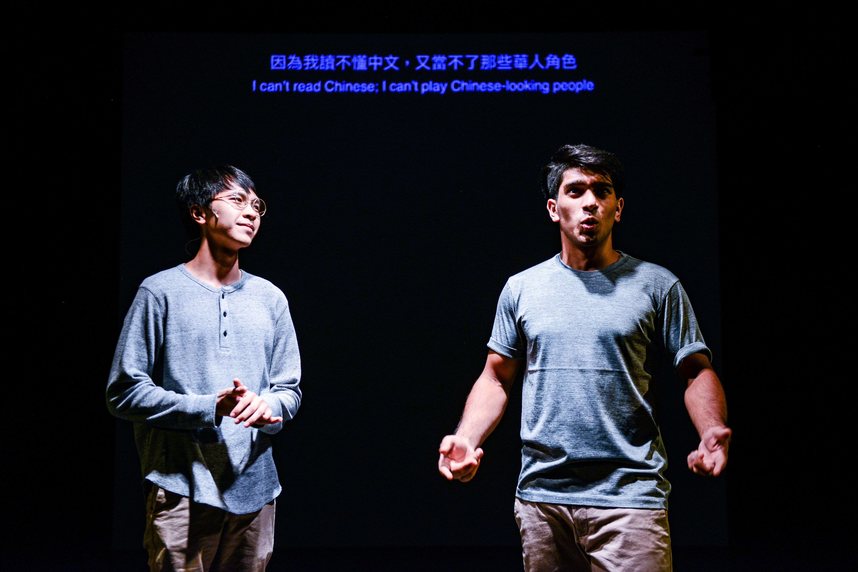 在天台製作（Rooftop Productions）的當代劇場作品《山下的證詞》演出中，演員巫加沙（Mohammad Kashif Ali）與吳景隆站在昏暗的舞台上，背景為純黑色。舞台上方投影出字幕：「因為我讀不懂中文，又當不了那些華人角色」，此幕展現了該劇探討文化認同與身份定位的深刻主題。