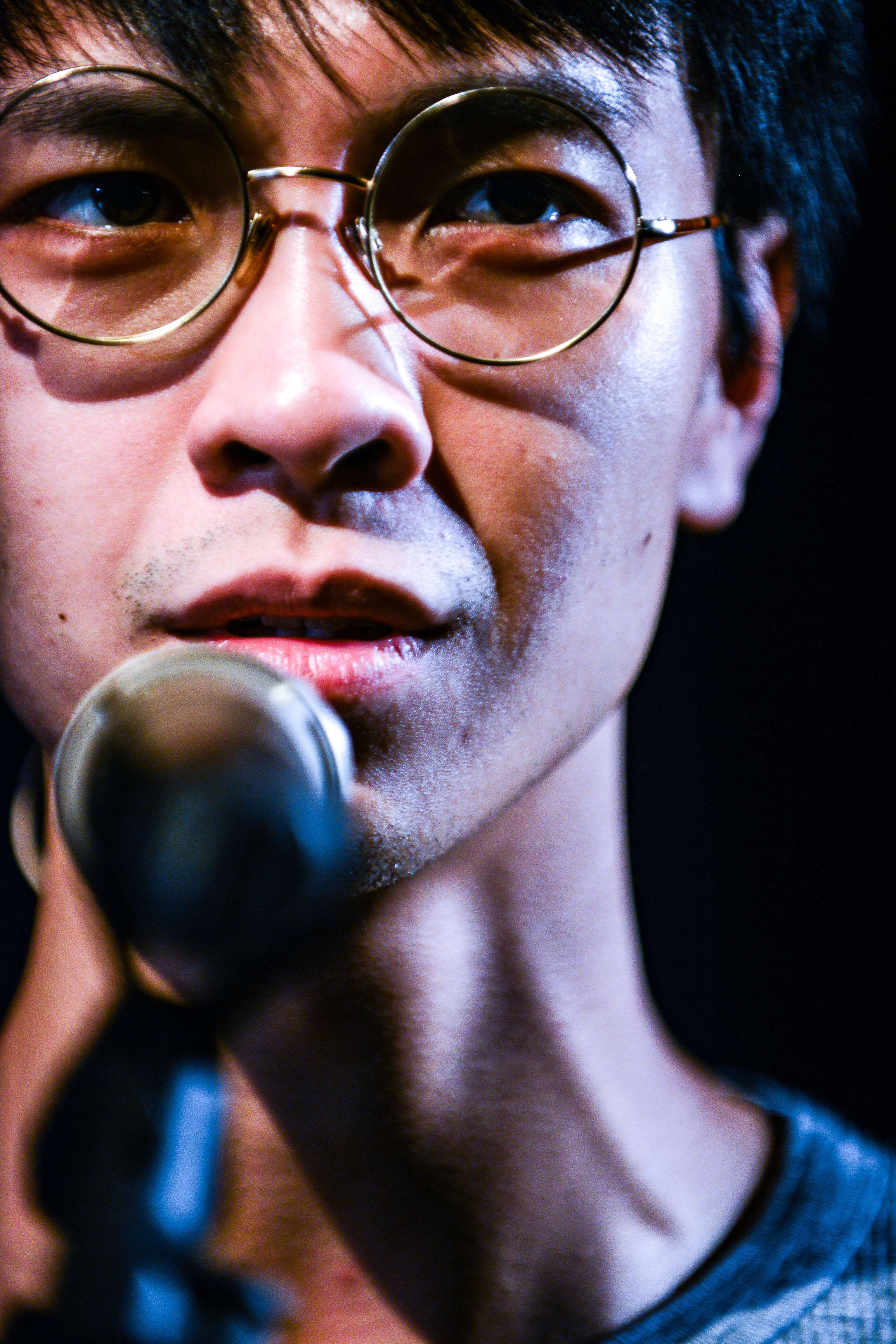 Close-up headshot of actor Ng King Lung during a performance of the contemporary Hong Kong theatre production 'Testimony' (《山下的證詞》). The stage lighting catches his intense, focused expression as he speaks into a microphone, highlighting his thin-rimmed round glasses and thoughtful gaze. He captures the essence of the character with the line: 'This isn’t my culture. But it’s OK, I’m a professional.'