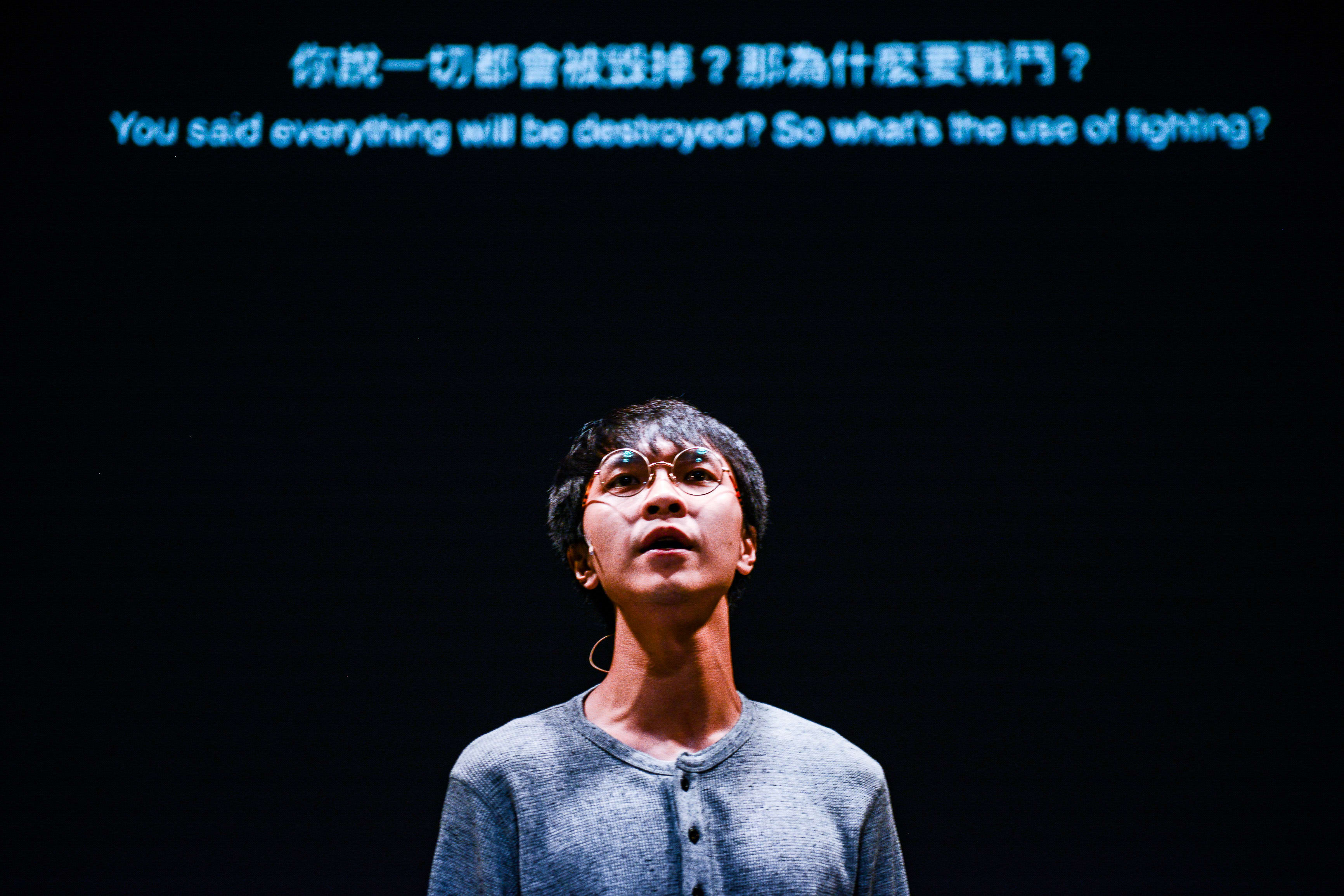 天台製作（Rooftop Productions）當代劇場作品《山下的證詞》（Testimony）的戲劇性舞台攝影。演員吳景隆在黑暗的背景中被單一聚光燈照亮，他佩戴圓框眼鏡，身穿休閒灰衫，神情深沉地仰望。在他身後的投影字幕寫道：「你說一切都會被毀掉？那為什麼要戰鬥？」