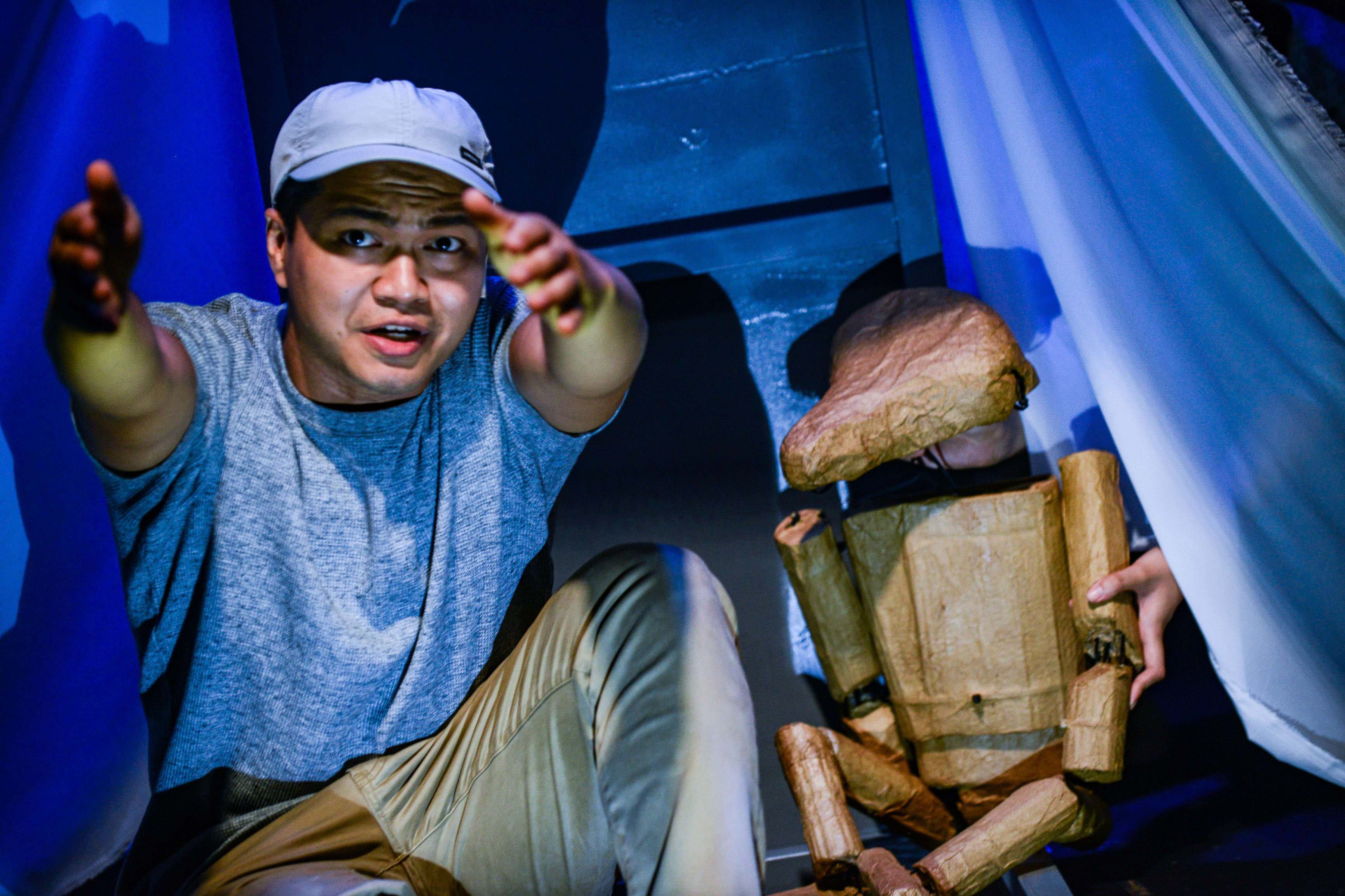 Basnet Tusar performs in a scene from Rooftop Productions’ contemporary play, 'Testimony'. He sits in a dimly lit, blue-toned stage setting, reaching toward the camera with an intense, earnest expression, while beside him, a crude, life-sized cardboard puppet sits partially obscured by a white curtain. As he interacts with the prop, he delivers the line, "We would stay out all night across the whole Chinese border."