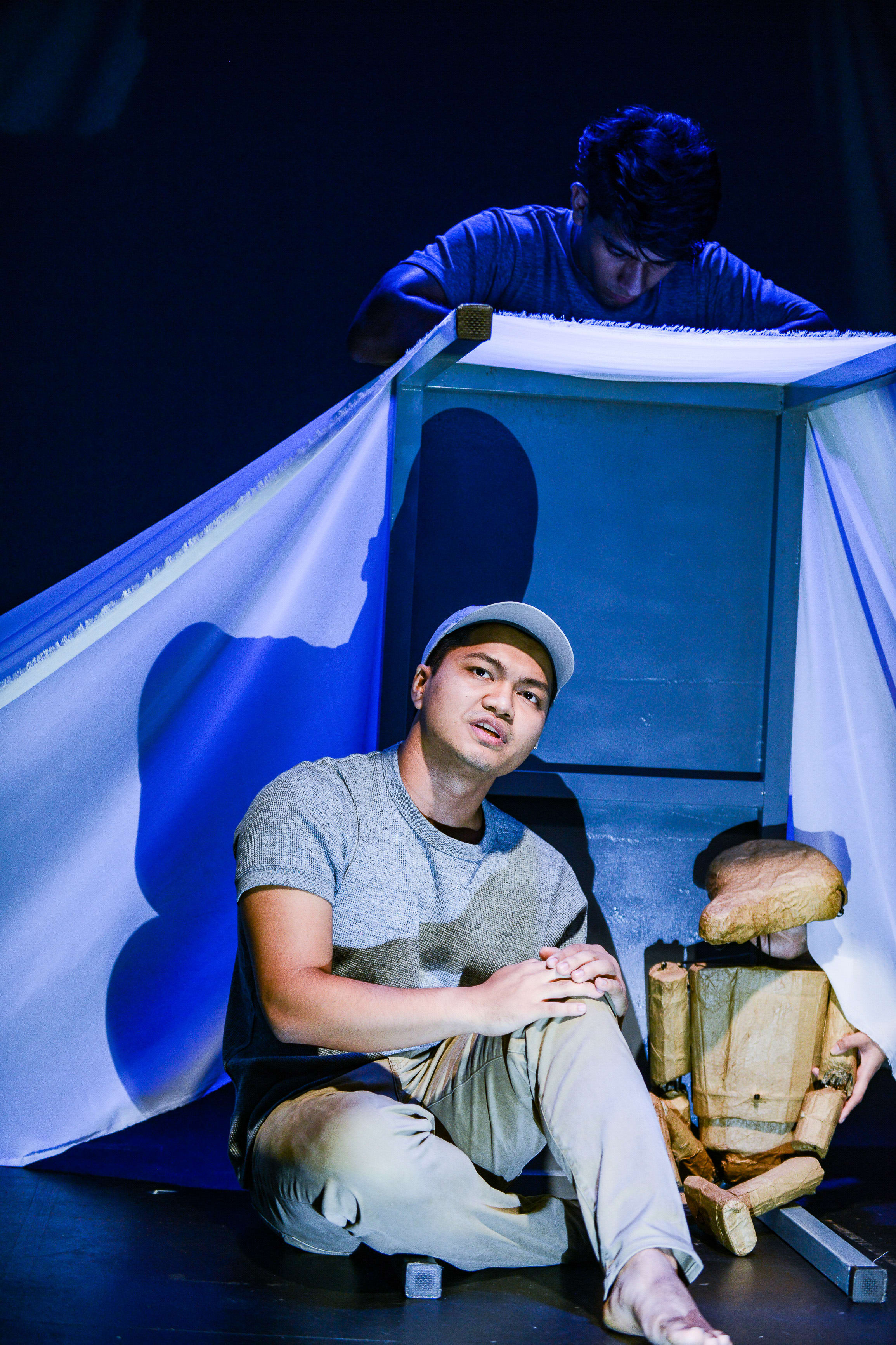 In a scene from Rooftop Productions' contemporary Hong Kong play Testimony, an actor wearing a grey t-shirt, beige trousers, and a white baseball cap sits on a dimly lit stage floor. He looks upward with a contemplative, slightly pained expression. Behind him, a second performer is partially visible, leaning over a square, table-like structure draped in a white cloth, while a rustic, wooden articulated puppet sits beside the seated actor. The lighting uses dramatic blue highlights, emphasizing the raw, experimental atmosphere typical of HK independent theatre. "Testimony."