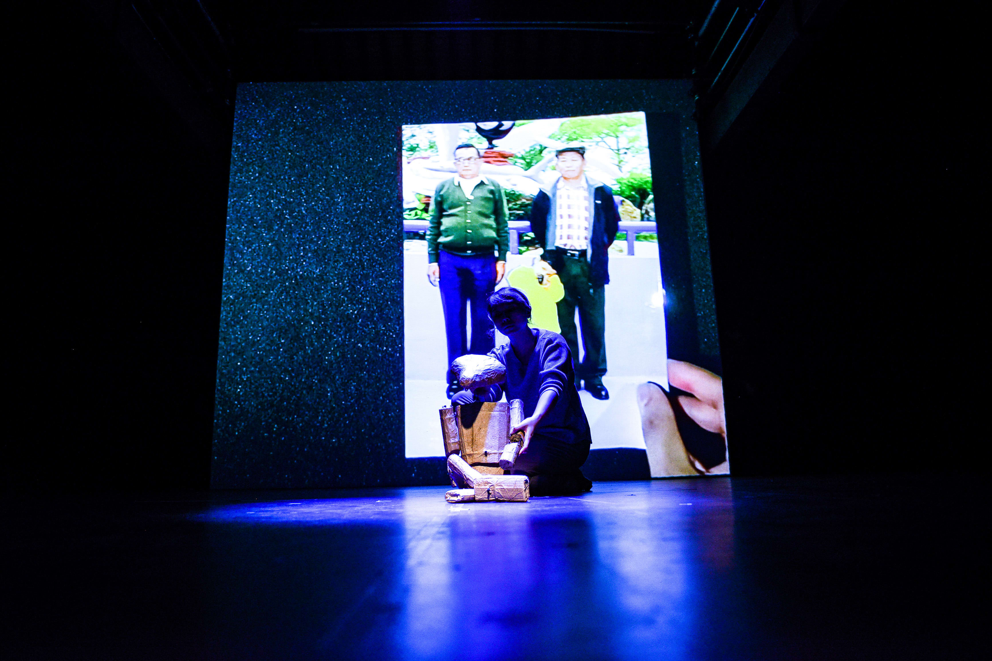 On a dimly lit stage, Arisa Yasaratne interacts with a wooden, robot-like puppet sculpture. Behind her, a large digital projection displays a vintage photograph of two men standing outdoors, providing a historical backdrop for this scene from Rooftop Productions' contemporary theatre piece, 'Testimony'. As she handles the puppet, she reflects on the past: 'My grandpa was stationed on the Hong Kong border from 1970 to 1976.'