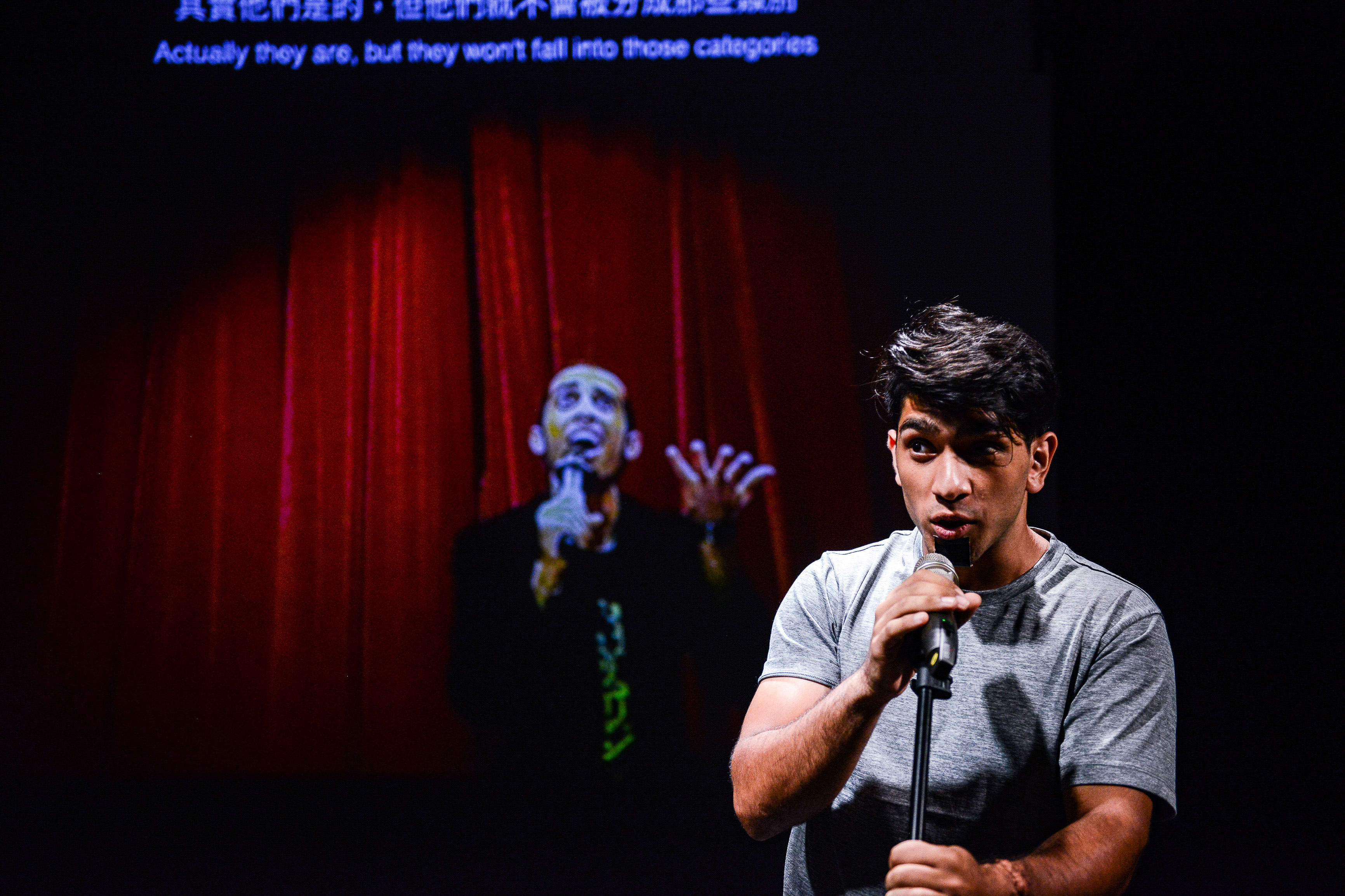A dramatic stage photo from Rooftop Productions' contemporary theatre piece 'Testimony' (《山下的證詞》). In the foreground, actor Mohammad Kashif Ali stands under a spotlight, holding a microphone and looking intensely toward the audience. Behind him, a large projection on a screen depicts a silhouette of a performer against a deep red curtain. The scene captures a powerful moment of reflection, as the actor speaks the line: 'I am in the middle, so I can see both worlds'.