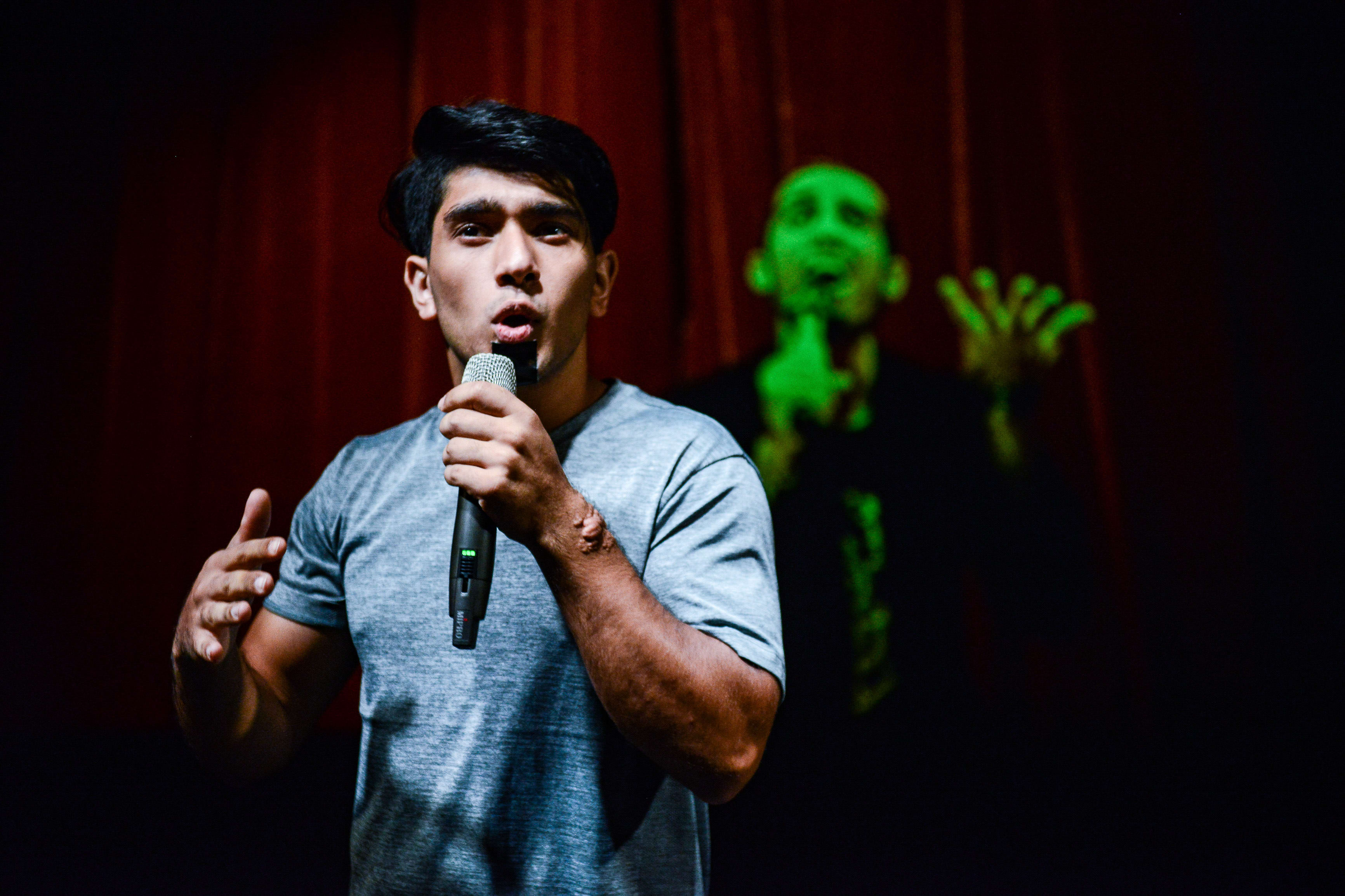In a dramatic scene from Rooftop Productions' contemporary theatre piece 'Testimony' (《山下的證詞》), actor Mohammad Kashif Ali stands center stage, illuminated by a spotlight while holding a microphone. He is dressed in a simple grey t-shirt, gesturing with his left hand as he delivers an intense, emotional monologue. In the darkened background, another performer is silhouetted and bathed in an eerie green light, emphasizing the production's evocative stagecraft. 'Testimony'
