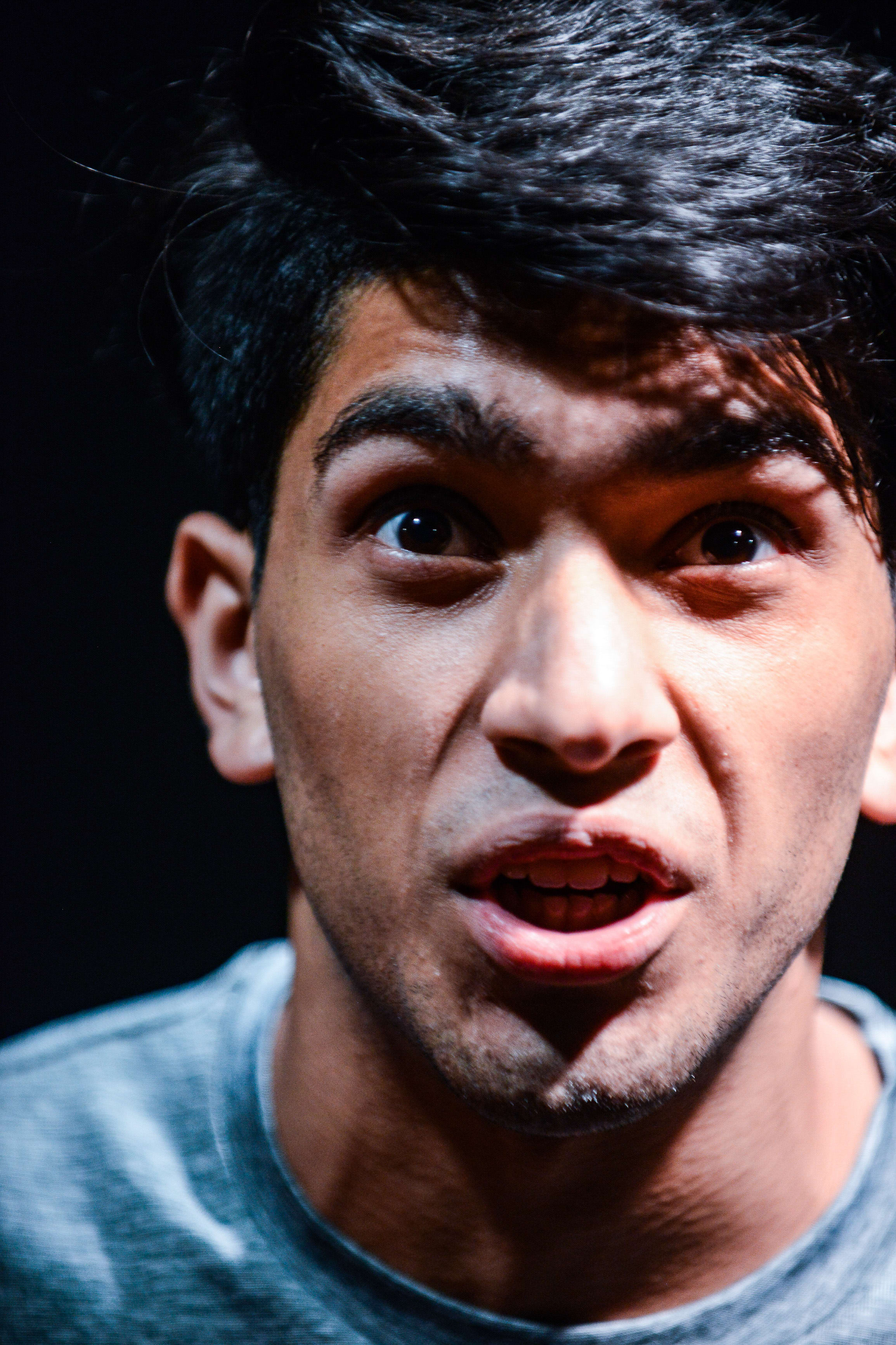 A dramatic close-up of actor Mohammad Kashif Ali during a rehearsal for the contemporary Hong Kong theatre production Testimony (《山下的證詞》). His expression is one of intense distress, with wide eyes and a slightly open mouth, illuminated by stark stage lighting that creates high-contrast shadows against a dark background. "Testimony"