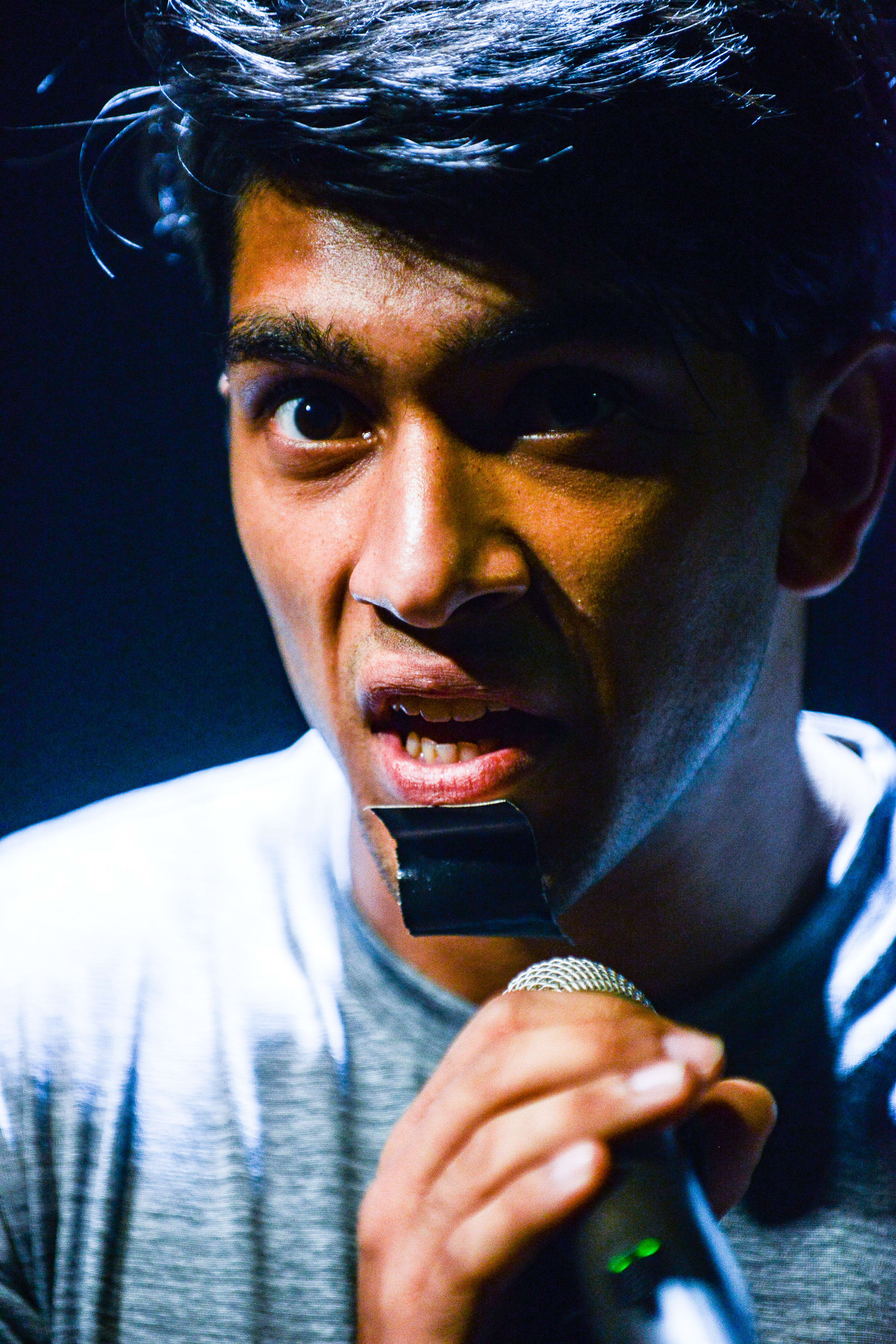 Close-up of actor Mohammad Kashif Ali performing in Rooftop Productions' contemporary theatre piece 'Testimony' (《山下的證詞》). Bathed in intense, dramatic stage lighting, he holds a microphone near his mouth, while a piece of black tape is visibly applied to his chin, adding a striking, raw aesthetic to the performance. His intense expression captures the heavy, emotional weight of the production. 'Testimony'