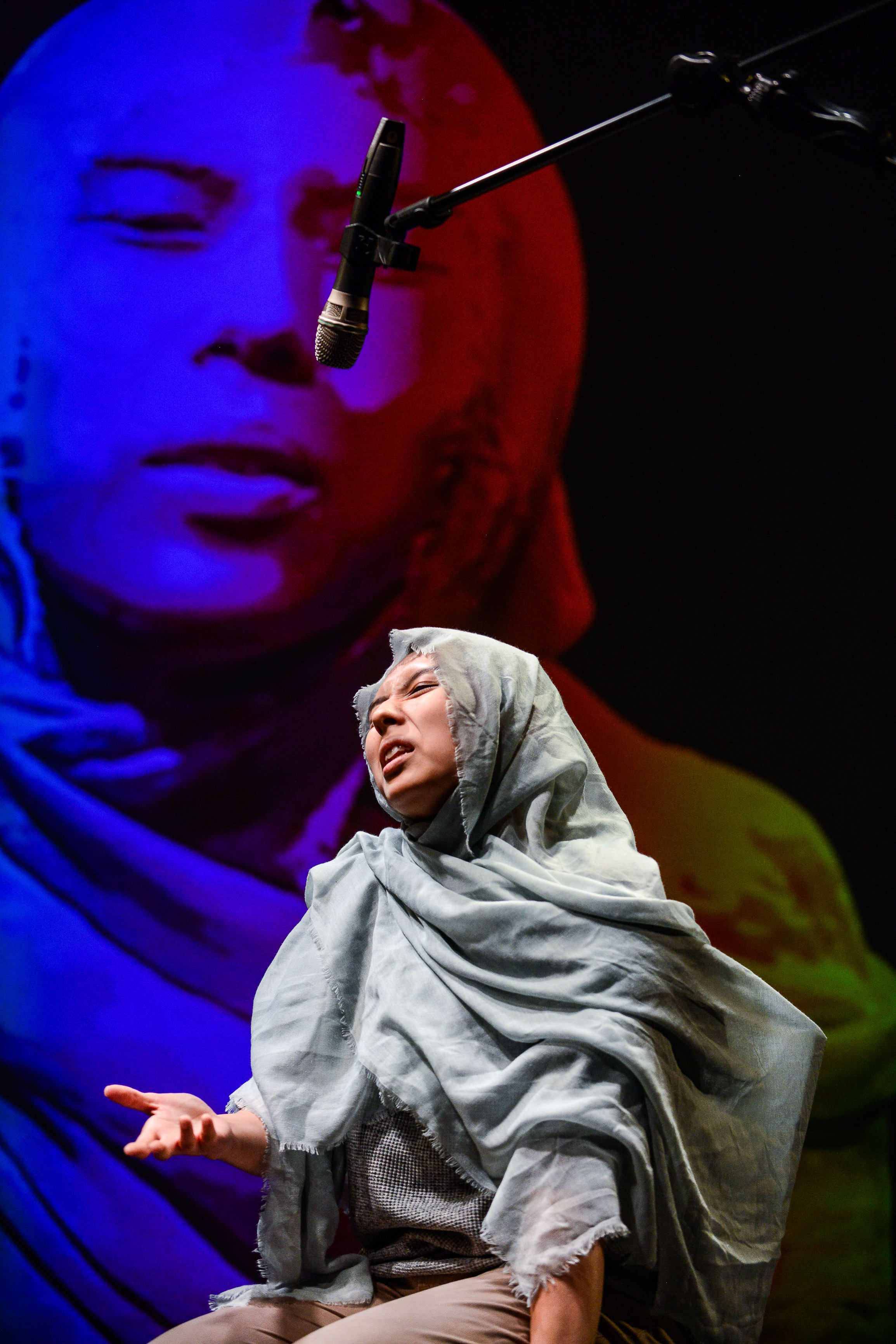 Arisa Yasaratne performs in Rooftop Productions' contemporary theatre piece 'Testimony' (《山下的證詞》). Bathed in intense blue and warm stage lighting, she wears a light-coloured headscarf and sits before a large, dramatic projection of a face, delivering an emotive monologue. As she speaks, she says, "You know, we need to do a little bit of acting."