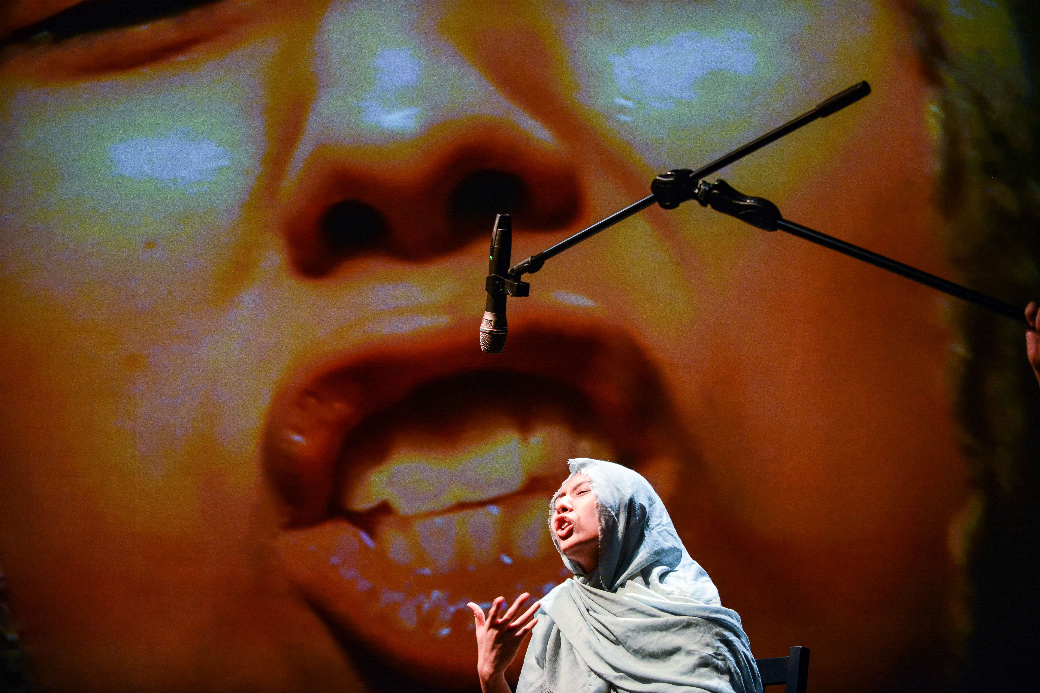 Performer Arisa Yasaratne stands on stage during a contemporary theatre performance of 'Testimony' (《山下的證詞》) by Rooftop Productions (天台製作), dressed in a light-colored headscarf and gesturing with an expressive hand. Behind her, a massive, out-of-focus projection of a human face dominates the stage backdrop, creating a powerful visual juxtaposition. As she engages with the multimedia stage design, she delivers a lines from the play, 'You, can you speak with an Indian accent?'