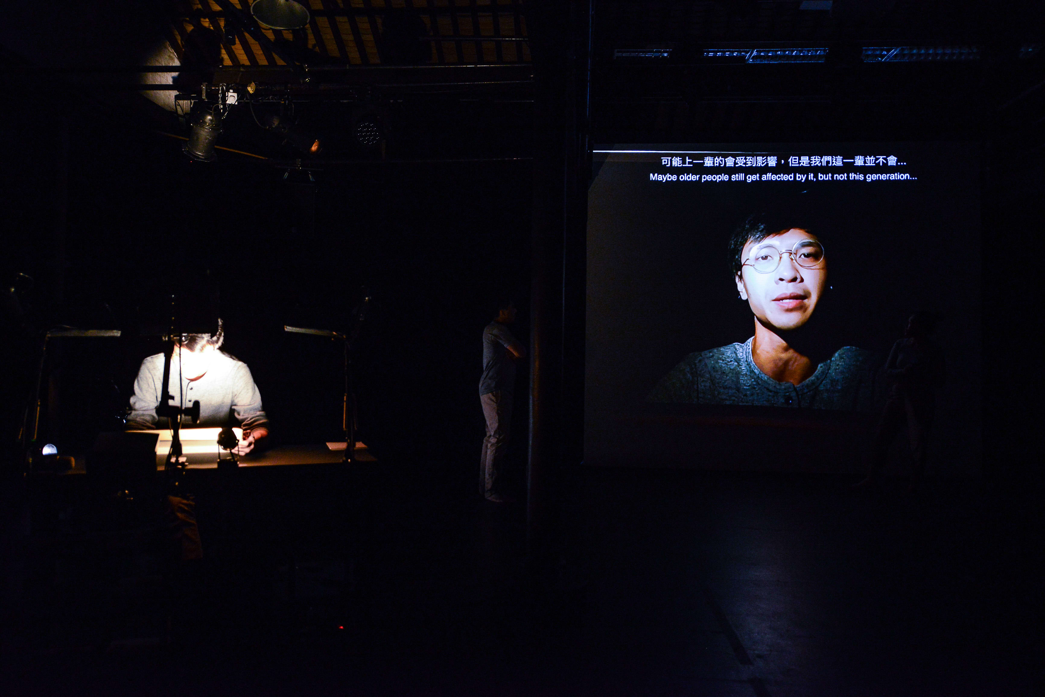 In a dim, atmospheric theatre setting, actor Ng King Lung sits at a desk under a focused spotlight while a large projection screen displays a close-up, dimly lit video portrait of him. This multimedia scene is from the Rooftop Productions (天台製作) performance, 'Testimony'. As Ng King Lung delivers a poignant monologue on stage, the screen shows him speaking the line: 'Maybe older people still get affected by it, but not this generation...'