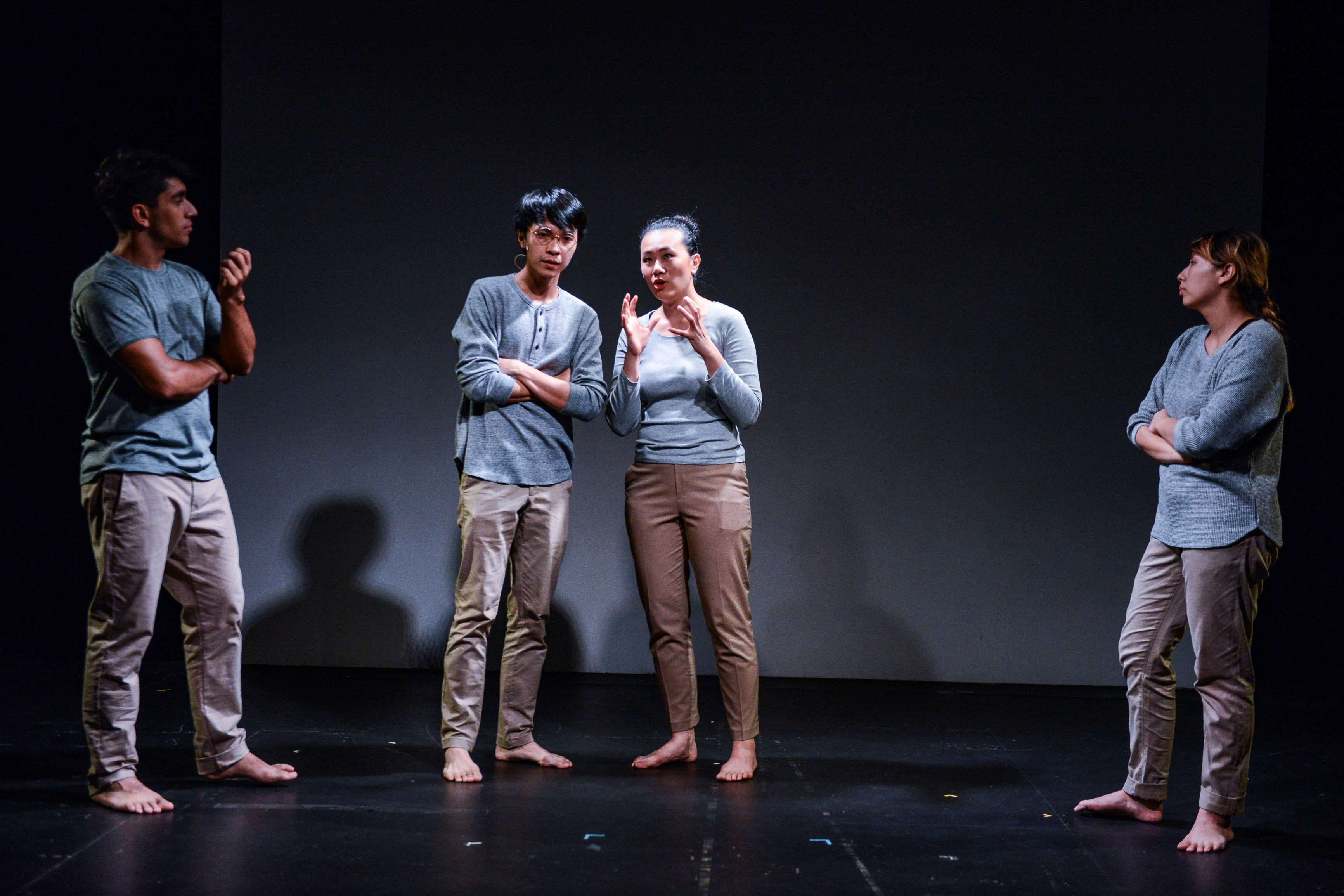 Four actors stand on a sparsely lit stage in a contemporary theatre performance of Rooftop Productions' (天台製作) show 'Testimony' (《山下的證詞》). From left to right: Mohammad Kashif Ali, Ng King Lung, Arisa Yasaratne, and Michelle Li are dressed in casual, earth-toned clothing. The actors are arranged in a line, with the central two engaged in a dramatic dialogue while the others look on with contemplative expressions, capturing the minimalist and poignant aesthetic of Hong Kong contemporary theatre. "Testimony"