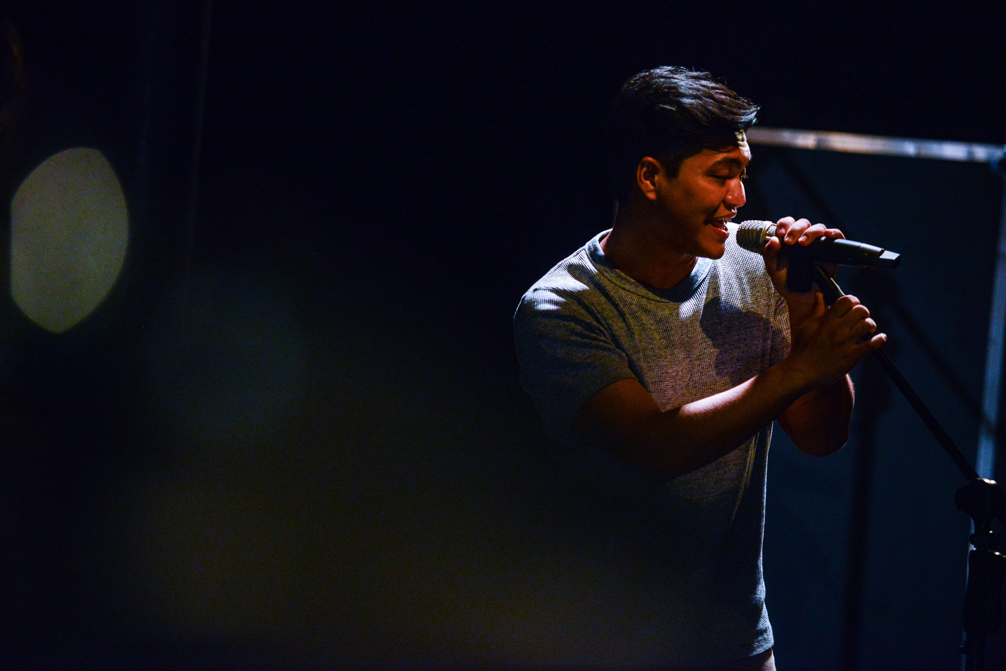 Basnet Tusar performs in a spotlight during a rehearsal for the contemporary Hong Kong theatre production Testimony. The actor is captured in profile, singing intensely into a microphone, with a dramatic, moody lighting setup typical of Rooftop Productions' immersive stage work. 'Testimony'
