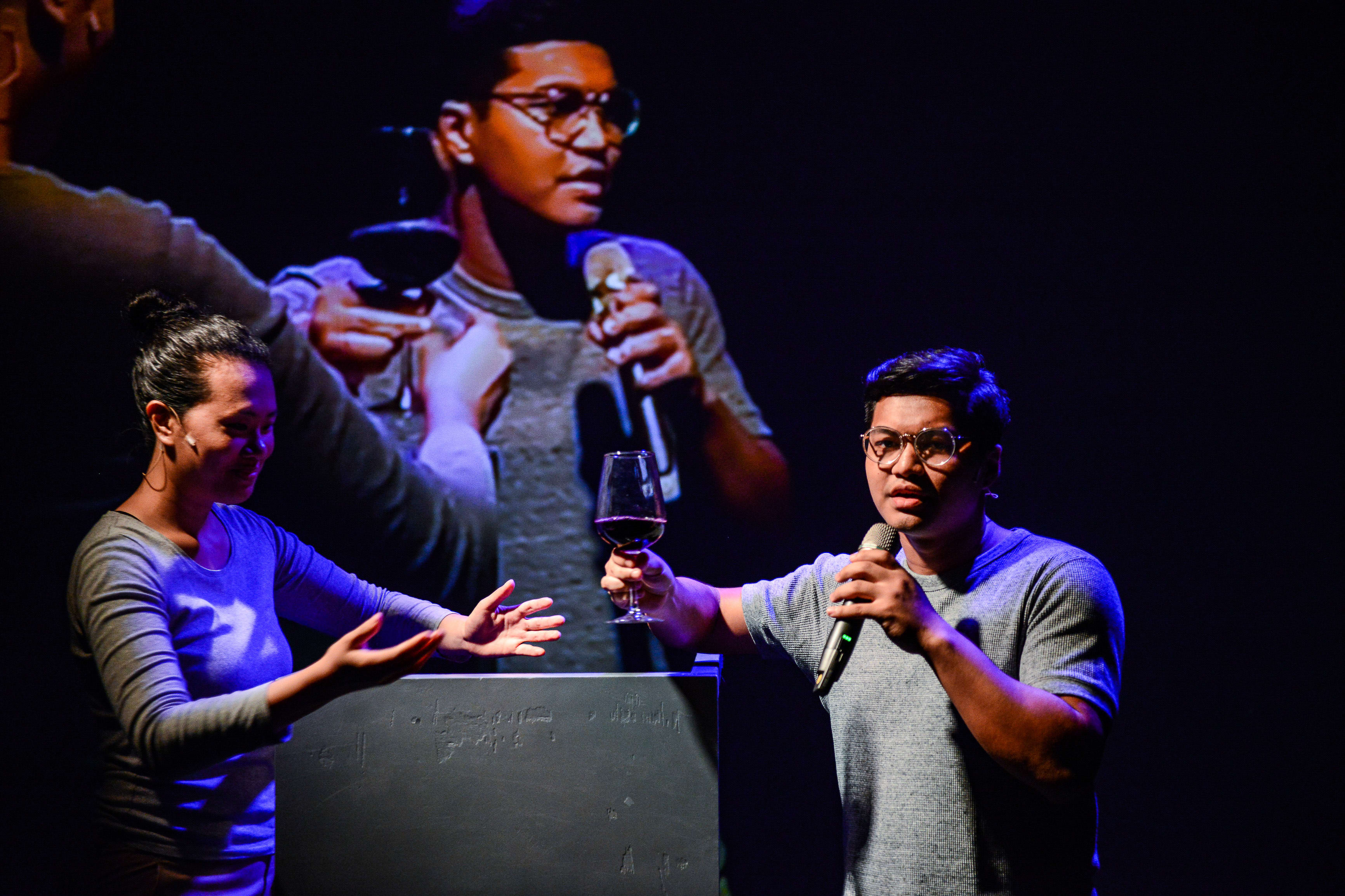 In this dramatic scene from Rooftop Productions' contemporary theatre piece 'Testimony' (《山下的證詞》), an actor wearing glasses holds a microphone and a glass of red wine, standing behind a lectern on a dimly lit stage. Beside him, a woman gestures with her hands as if speaking, while a large, ghostly projection of the same man looms in the background, creating a surreal, reflective atmosphere. This performance explores themes of memory and truth, characteristic of Rooftop Productions' unique artistic style. As the actor delivers his lines, he reflects: 'Testimony.'