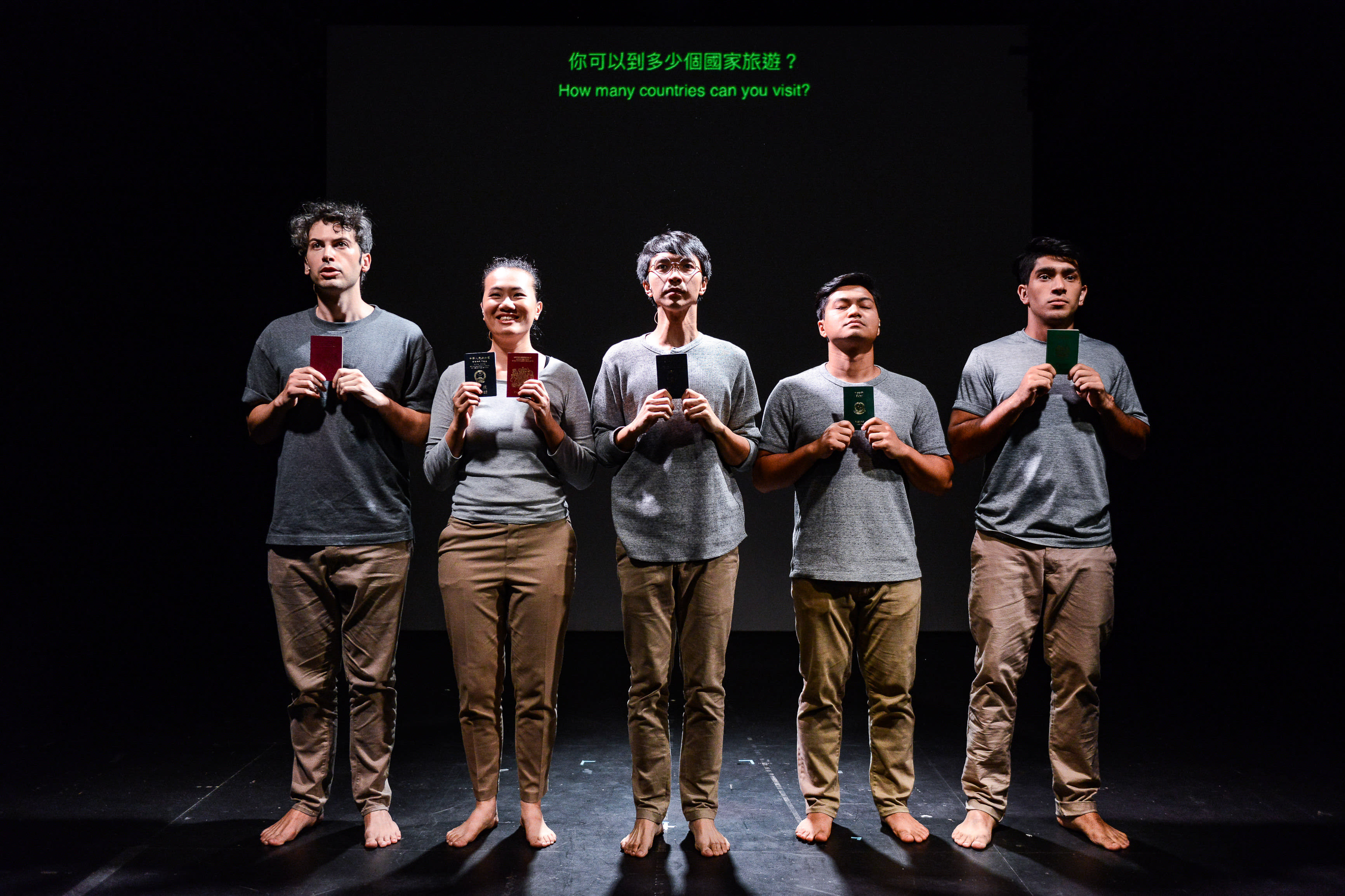 In a dramatic scene from Rooftop Productions' contemporary theatre piece 'Testimony', five actors stand in a line on a dimly lit, minimalist stage against a black backdrop. Each performer holds a single passport, representing varying levels of global mobility. The actors are, from left to right: Ivor Houlker, Michelle Li, Ng King Lung, Mohammad Kashif Ali, and Basnet Tusar. Above them, a digital screen displays the question 'How many countries can you visit?'. This powerful stage production explores themes of privilege, citizenship, and identity in Hong Kong. The performers stand solemnly as they reenact the line, 'Line up in order of how many countries you can visit without a visa.'