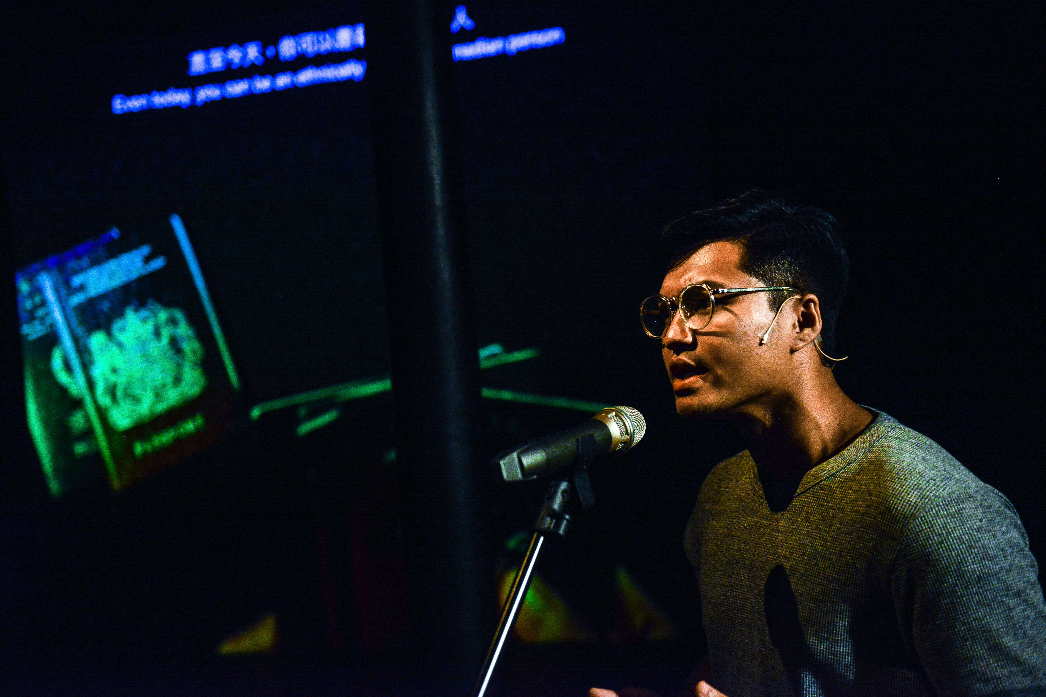 在天台製作（Rooftop Productions）的當代劇場作品《山下的證詞》（Testimony）中，演員 Basnet Tusar 在低調的舞台燈光下神情專注地對著麥克風進行演出。背景投影著護照的綠色光影，營造出強烈的戲劇氛圍。《山下的證詞》
