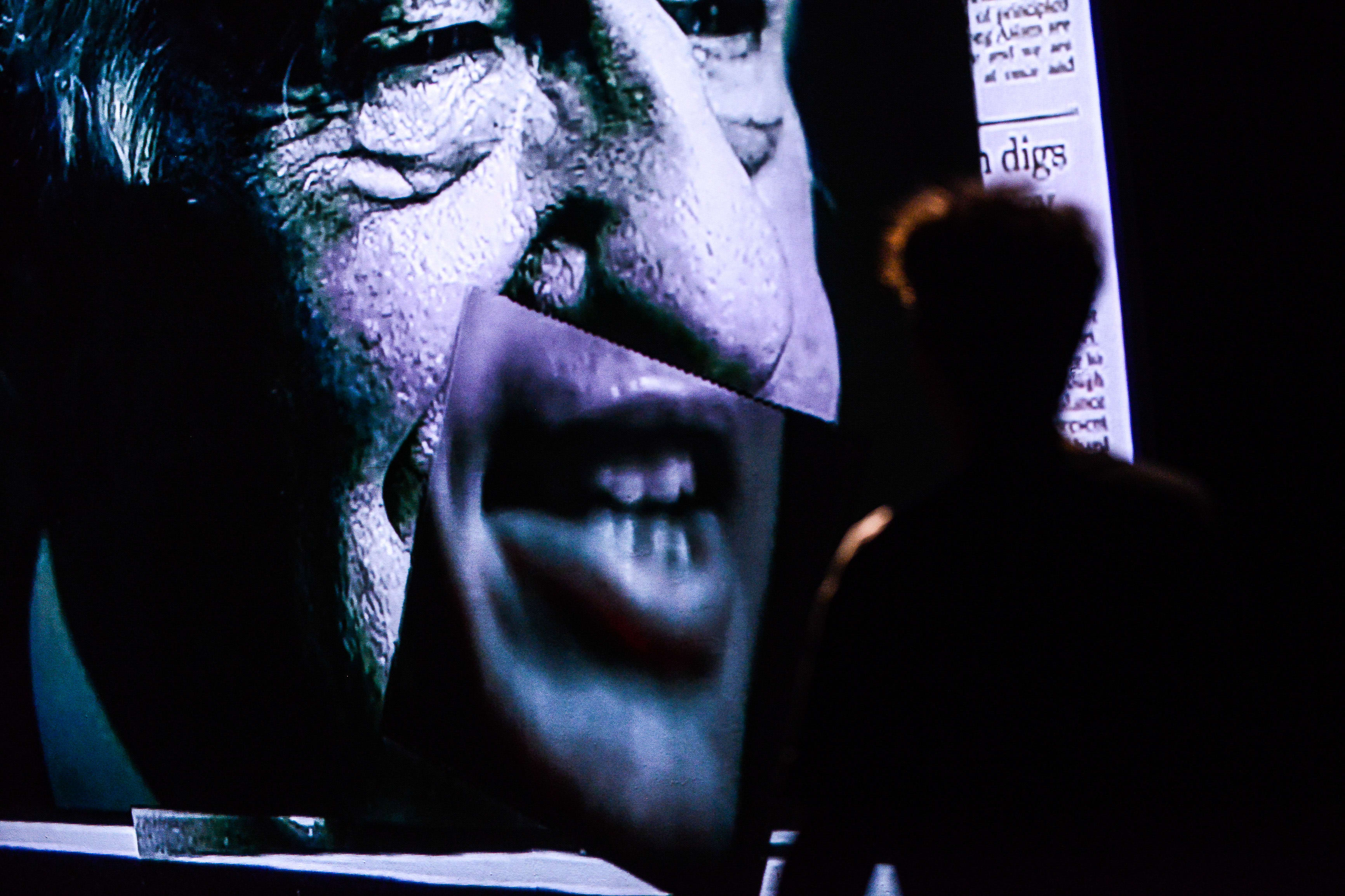 In a dramatic scene from Rooftop Productions' contemporary theatre piece 'Testimony' (《山下的證詞》), a performer, Ivor Houlker, stands in silhouette before a large, high-contrast projection. The screen displays a close-up of a weathered, aging face, with a smaller, surreal insert of a mouth articulating speech, set against a dark background reminiscent of a political debate. The lighting emphasizes the stark, haunting atmosphere characteristic of the production. As the performer speaks to the audience, the projection echoes the line: 'And David Wilson came down from Scotland to participate in the debate.'