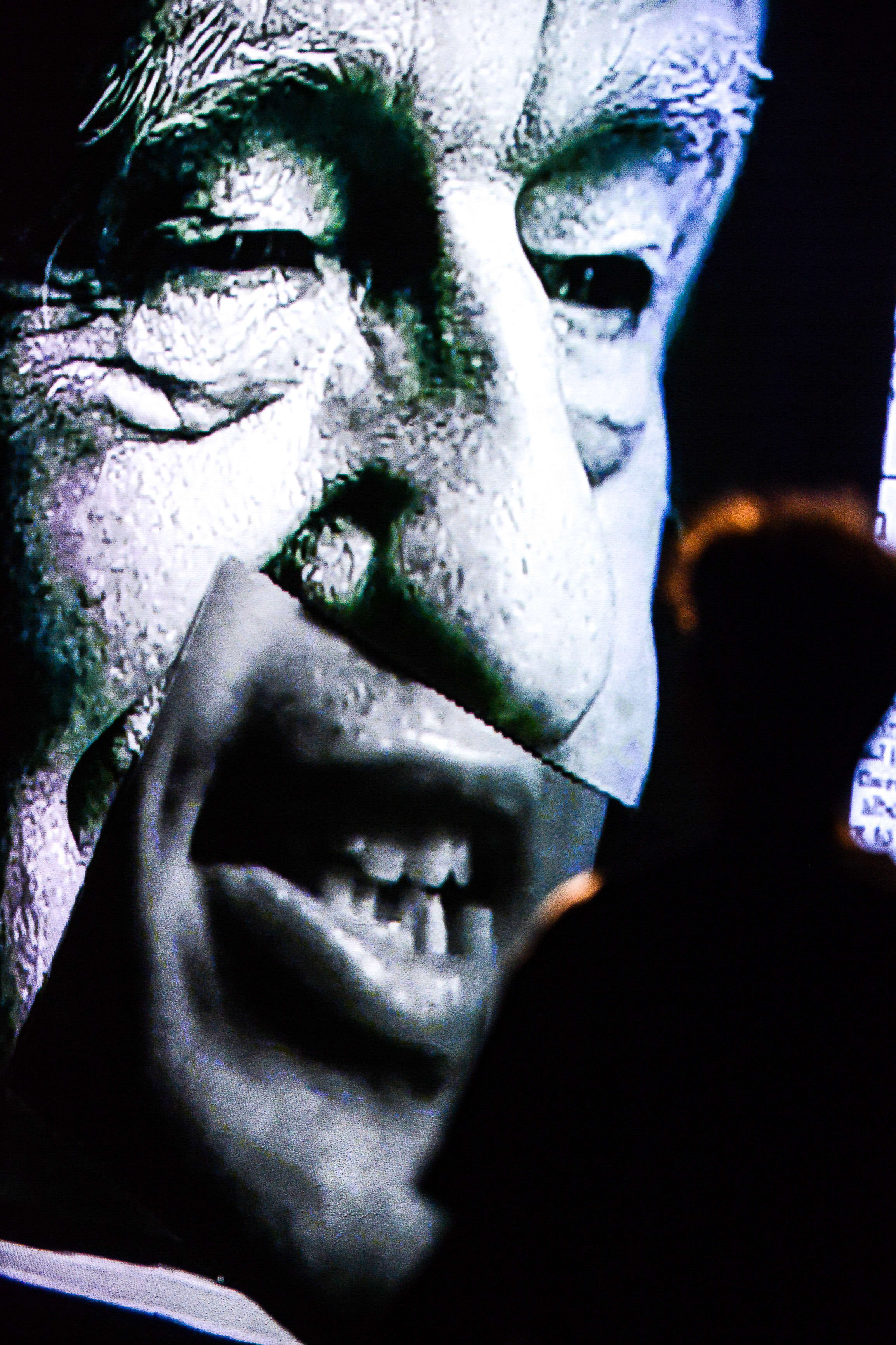 A dramatic close-up shot from the contemporary Hong Kong theatre production 'Testimony' (《山下的證詞》), featuring a larger-than-life, high-contrast projection of actor Ivor Houlker. The projection captures a detailed, intense facial expression, while the silhouette of an audience member or performer is visible in the darkened foreground, emphasizing the immersive staging typical of Rooftop Productions (天台製作). The lighting creates a stark, haunting atmosphere on stage. As the actor delivers his lines, he says: 'Testimony'