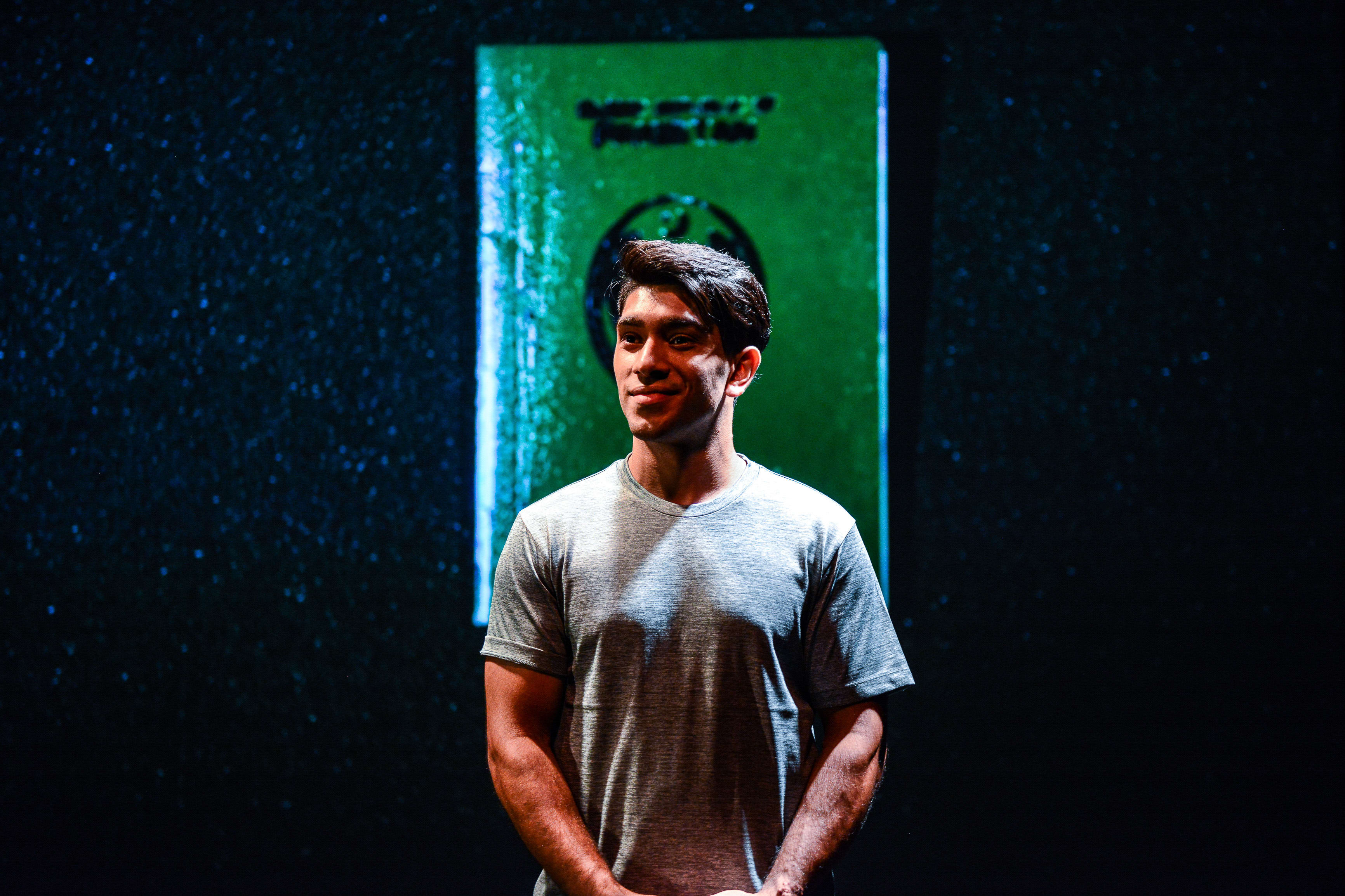 A dramatic stage shot from Rooftop Productions' contemporary theatre piece 'Testimony' (山下的證詞). Actor Mohammad Kashif Ali stands center stage, illuminated by a spotlight against a dark, textured background. He wears a simple grey t-shirt and looks slightly off-camera with a contemplative, serene expression. Directly behind him hangs a prominent vertical green banner featuring a circular logo, adding a bold visual element to the performance. 'Testimony'