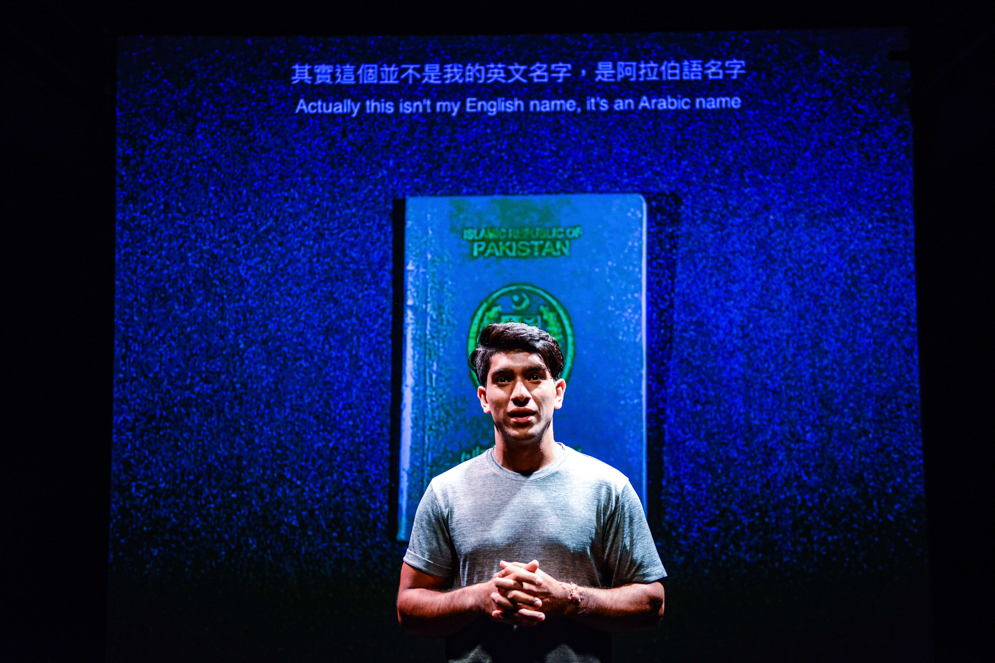 A dramatic stage shot from Rooftop Productions' contemporary theatre piece 'Testimony' ('山下的證詞'). The performer Mohammad Kashif Ali stands center stage, illuminated by soft lighting against a deep blue backdrop featuring a large, glowing projection of a Pakistani passport. He gestures calmly while speaking, his expression earnest and contemplative. As the performer speaks to the audience, he says, 'Actually this isn’t my English name, it’s an Arabic name.'