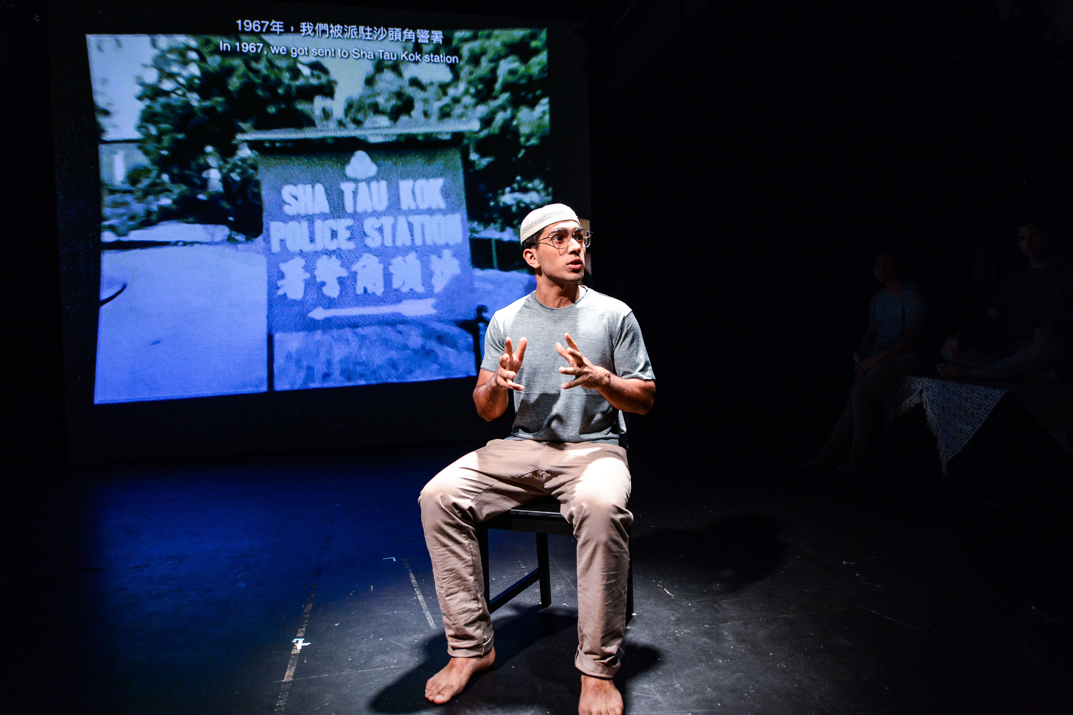In a dramatic scene from Rooftop Productions' contemporary theatre piece 'Testimony' (《山下的證詞》), actor Mohammad Kashif Ali sits on a simple wooden chair center-stage, illuminated by a stark spotlight in an otherwise dark, minimalist theatre space. He wears a grey t-shirt and light-colored trousers, gesturing expressively with his hands while narrating a historical account. Behind him, a large projection displays a vintage, high-contrast photograph of the 'Sha Tau Kok Police Station' with text referencing the events of 1967. As the performer reflects on this historical moment in the play, he recounts, '1000 people came to Sha Tau Kok village, came shouting, along the border.'