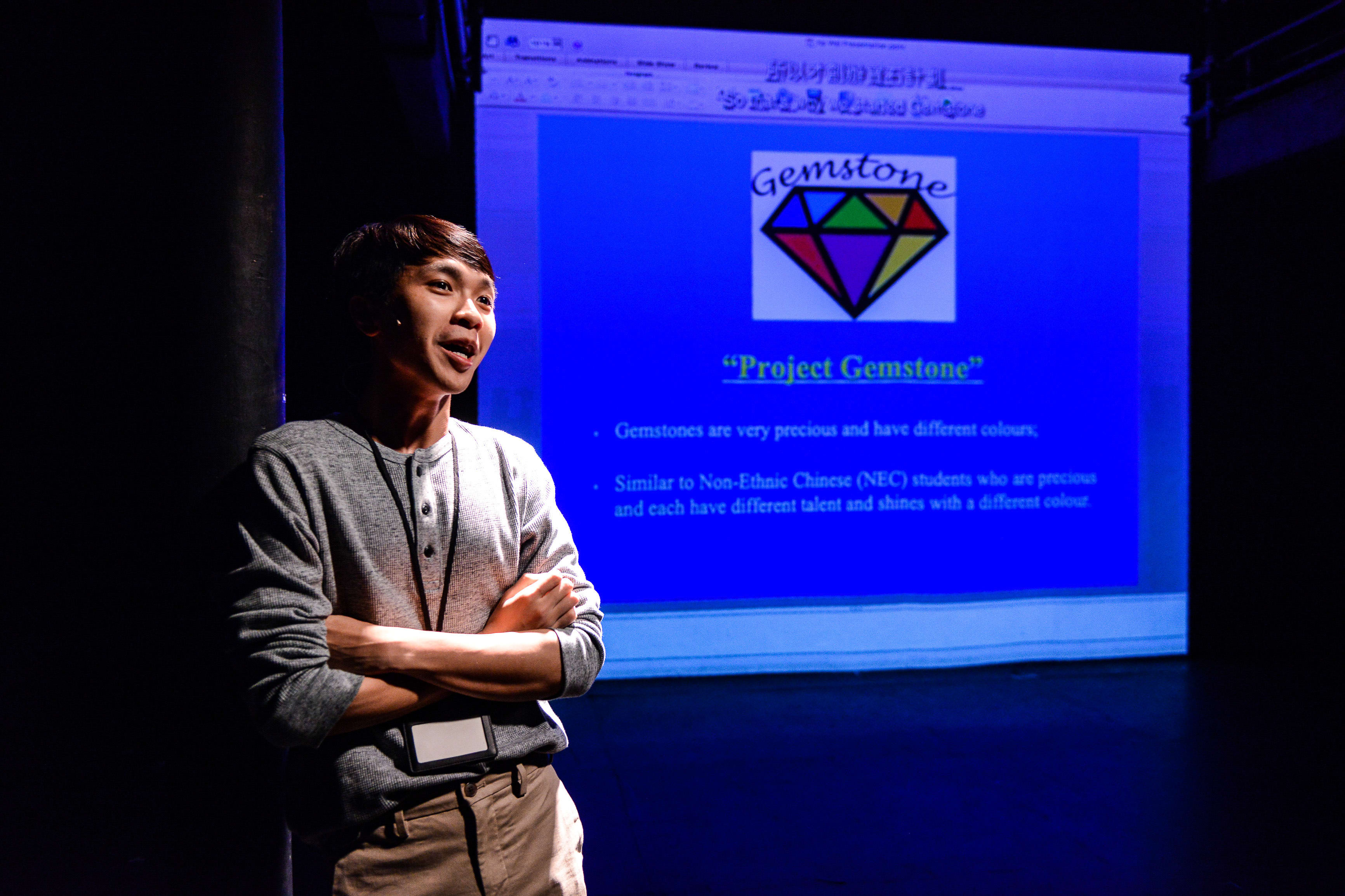 在天台製作（Rooftop Productions）的戲劇作品《山下的證詞》（Testimony）中，演員吳景隆站在舞台上，雙臂交叉，在黑暗的背景中被聚光燈照亮。他身後的投影幕顯示著名為「Project Gemstone」的演示文稿，圖中印有一個彩色鑽石圖案，文字將寶石的價值與非華裔（NEC）學生的多元才華作出了比喻。《山下的證詞》