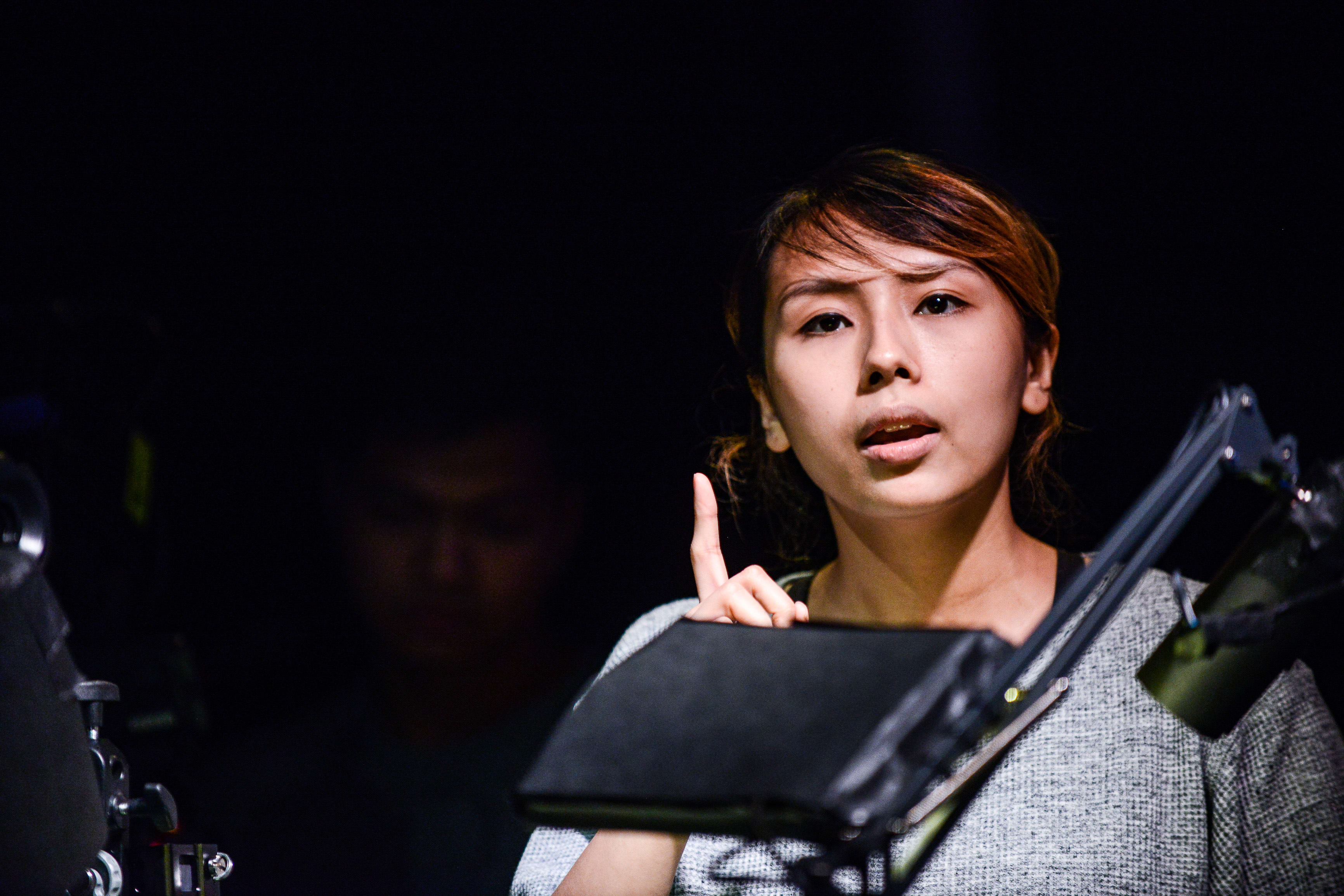 Arisa Yasaratne performs in a rehearsal for the contemporary Hong Kong theatre production 'Testimony' (《山下的證詞》), illuminated by dramatic stage lighting against a dark background. She gestures with her index finger raised, capturing an intense, thoughtful moment. As she delivers her lines during this scene, she remarks, 'There as a movie recently about the world’s most famous Parsee.'