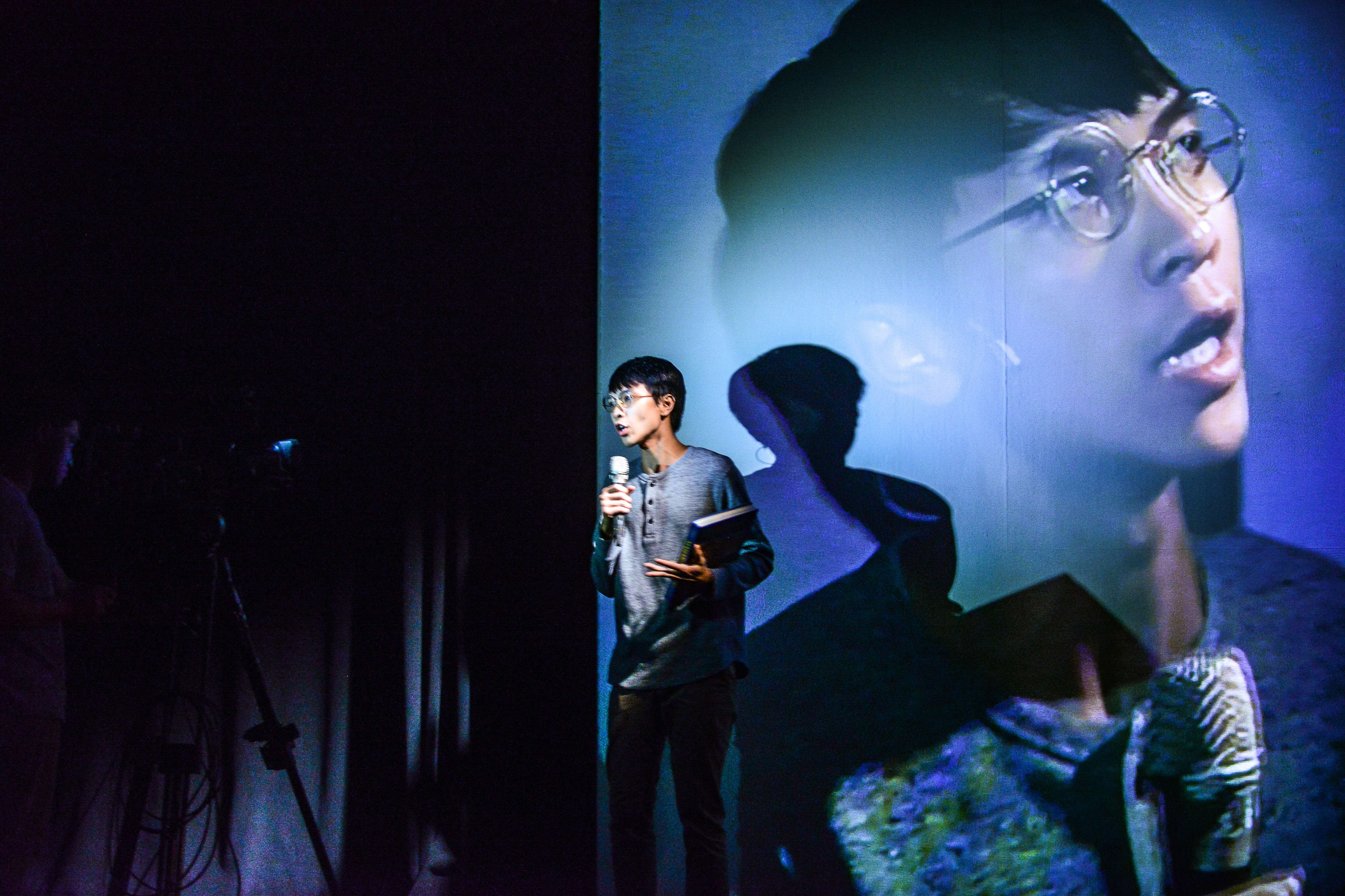 On a dimly lit stage, actor Ng King Lung stands holding a microphone and a folder, delivering a poignant performance in Rooftop Productions' contemporary theatre piece, 'Testimony'. A large-scale projection of Ng's face looms behind him, creating a surreal, double-exposure effect that emphasizes the themes of memory and confession. The dramatic blue lighting highlights his intense expression as he presents his 'Testimony'.