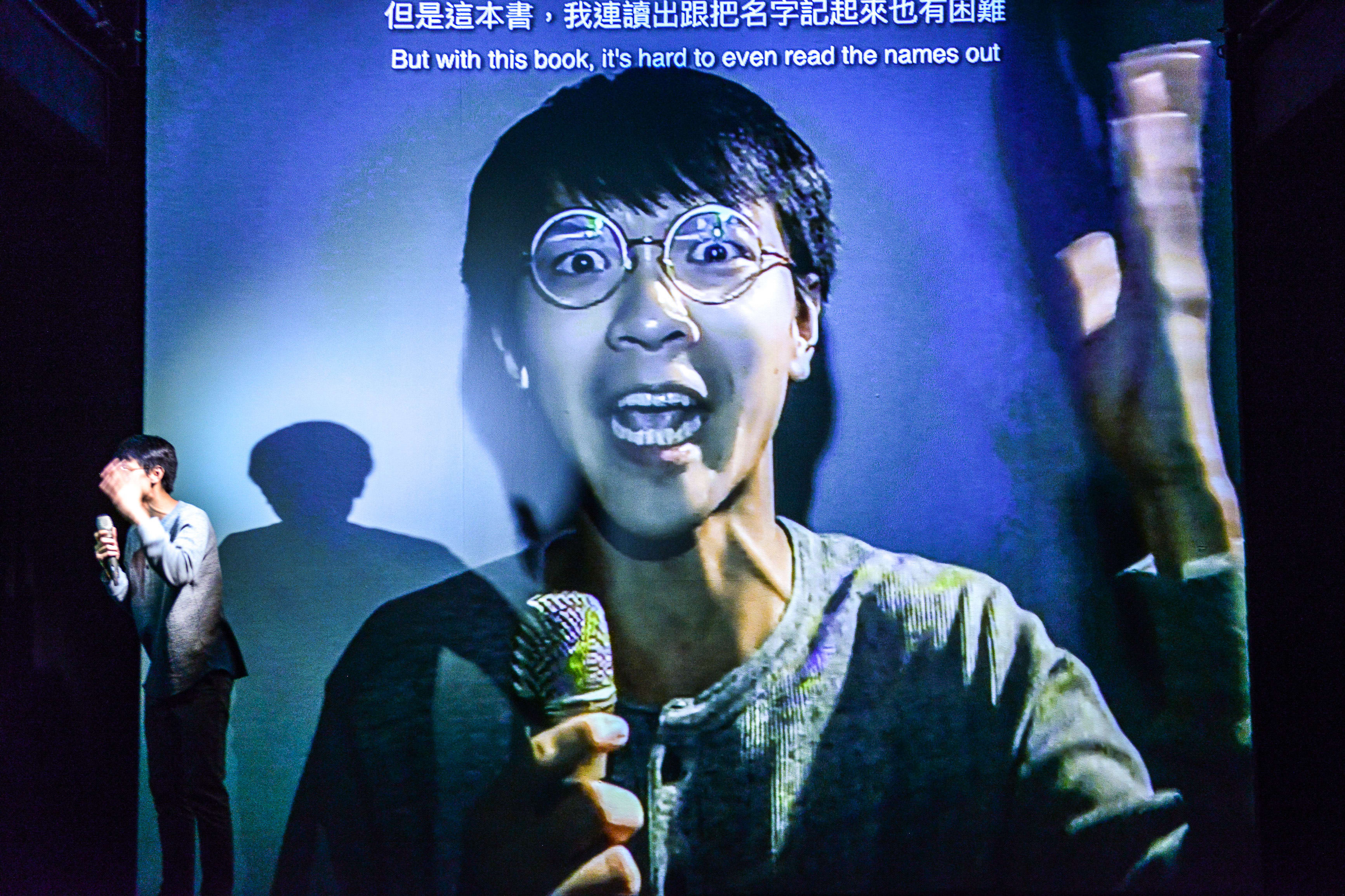 In a multimedia performance of the Rooftop Productions (天台製作) show 'Testimony' (《山下的證詞》), actor Ng King Lung stands on a dark stage, his figure partially illuminated by a cool, blue-toned projection behind him. The projection features a larger-than-life, high-contrast close-up of Ng King Lung wearing round glasses and speaking into a microphone with an expressive, animated face. The theatrical lighting highlights the tension between the live performer and his digital persona. As he interacts with the multimedia display, Ng King Lung delivers a monologue: 'I'm pretty familiar with Athenian democracy, the Renaissance, Qin Shi Huang. But with this book, it's hard to even read the names out.'