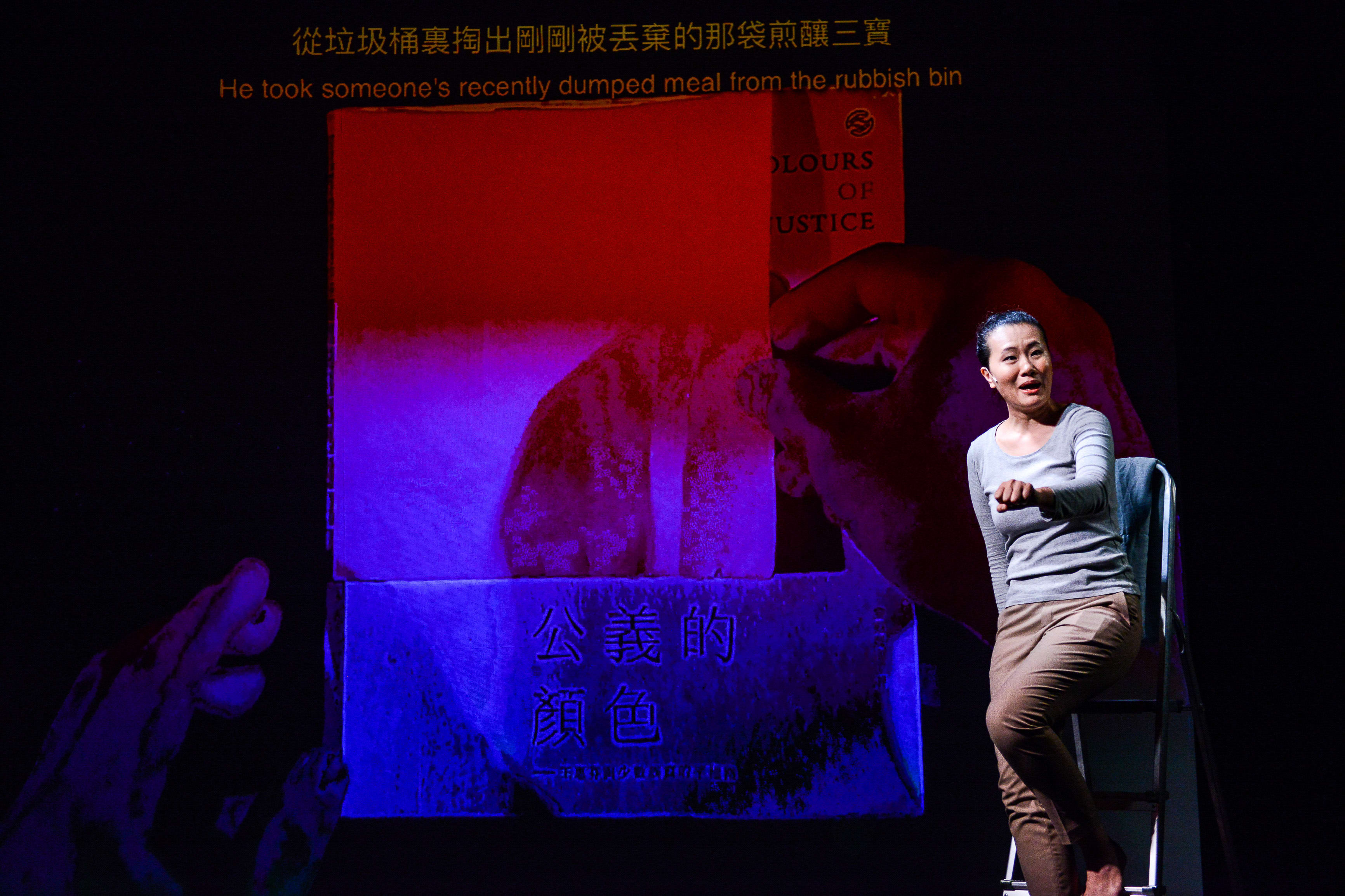 In a dramatic scene from Rooftop Productions' contemporary theatre piece 'Testimony' (《山下的證詞》), actress Michelle Li sits on a small ladder against a dark stage backdrop. A large, stylized projection of a human hand reaches toward a vivid red screen featuring the text 'Colours of Justice'. The lighting casts deep blue and purple hues, underscoring the raw, emotive storytelling typical of Hong Kong independent theatre. Michelle Li portrays the intensity of the narrative as she delivers the line: "He took someone's recently dumped meal from the rubbish bin."