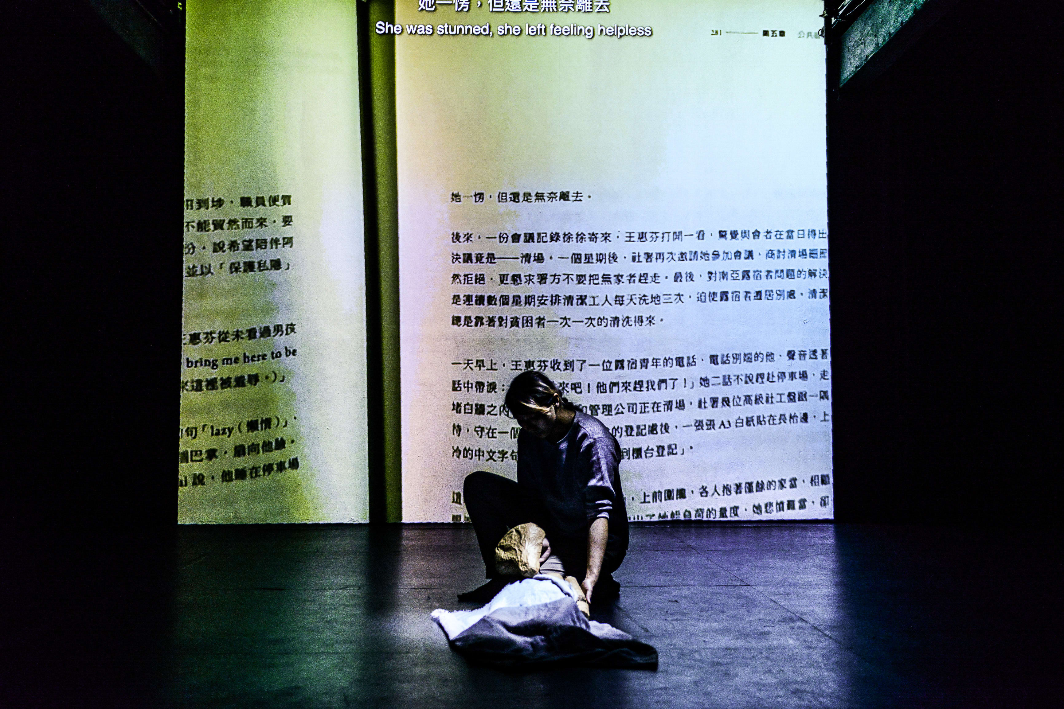 在天台製作 (Rooftop Productions) 的當代劇場作品《山下的證詞》(Testimony) 中，一位演員於昏暗的舞台上跪地，身旁覆蓋著一具用布遮蓋的形體，背景投映著大幅的文獻文字，呈現出強烈的歷史沉重感與當代劇場的敘事張力。這一幕展現了演出中深刻的情感張力與舞台美學。劇中有一句對白：「牆上面，用紅色的筆寫著『沒有將來』。」