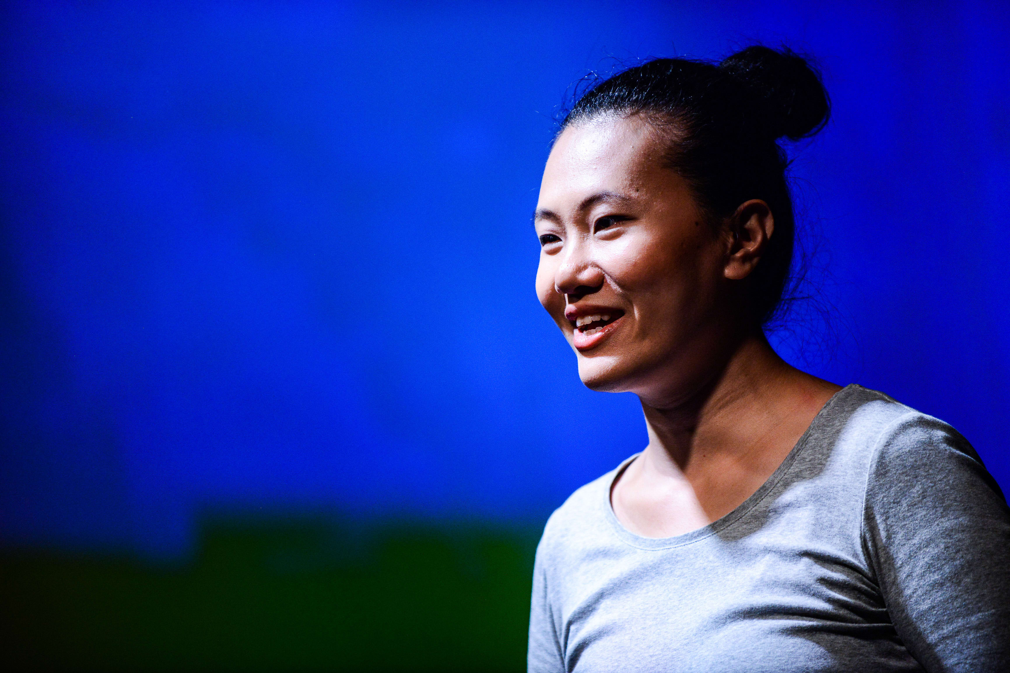 A close-up photograph of actor Michelle Li performing in the contemporary Hong Kong theatre production Testimony (《山下的證詞》) by Rooftop Productions. She is lit by a striking blue stage light, wearing a simple grey top with her hair in a bun, captured with a thoughtful, emotive expression that highlights the intimate nature of the performance. 'Testimony'