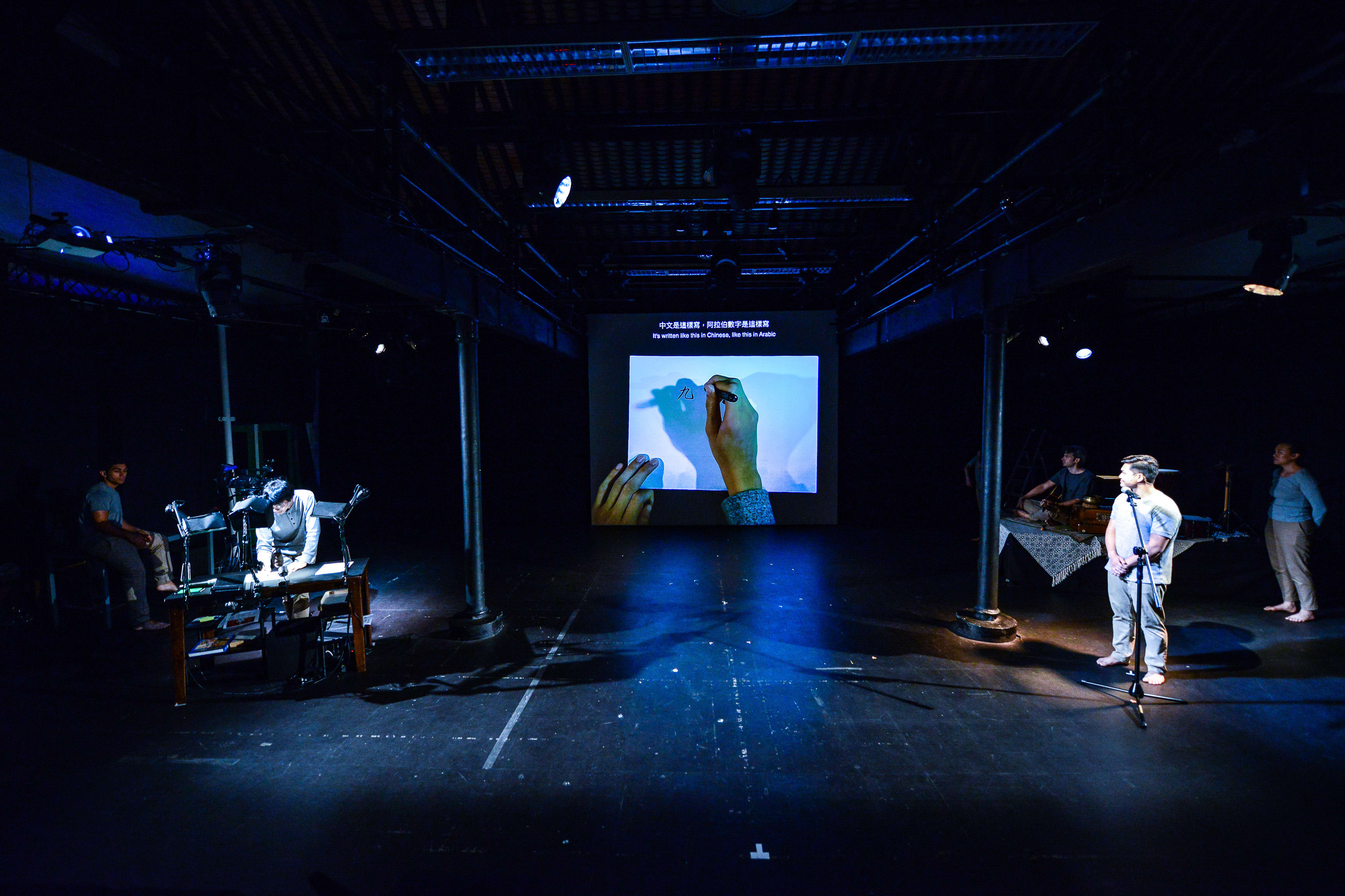 A wide shot of a contemporary theatre performance by Rooftop Productions (天台製作) titled 'Testimony' (山下的證詞). On a darkened stage, an actor stands stage right under a spotlight, speaking into a microphone, while another performer sits at a desk stage left, creating live ink drawings projected onto a large central screen. The production reflects the minimalist, experimental aesthetic of Hong Kong contemporary theatre. 'Testimony'