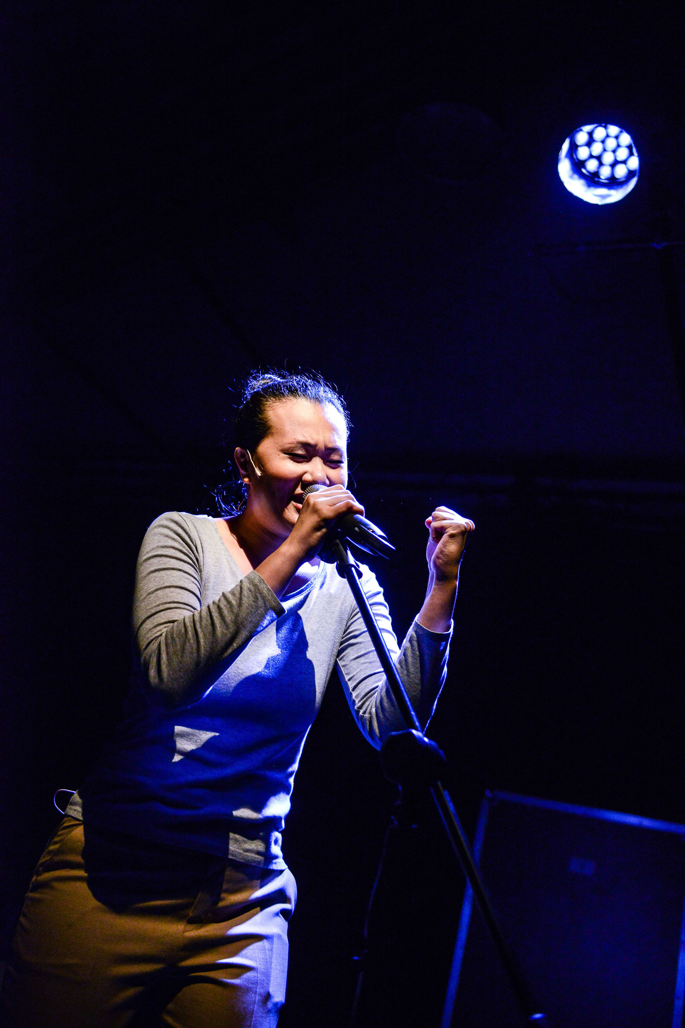 A performer, Michelle Li, passionately singing into a microphone on stage during a performance of the contemporary Hong Kong theatre production Testimony (《山下的證詞》). Bathed in a dramatic blue spotlight, she clenches her left hand in an emotive gesture, capturing a powerful moment of theatrical storytelling. "Testimony"